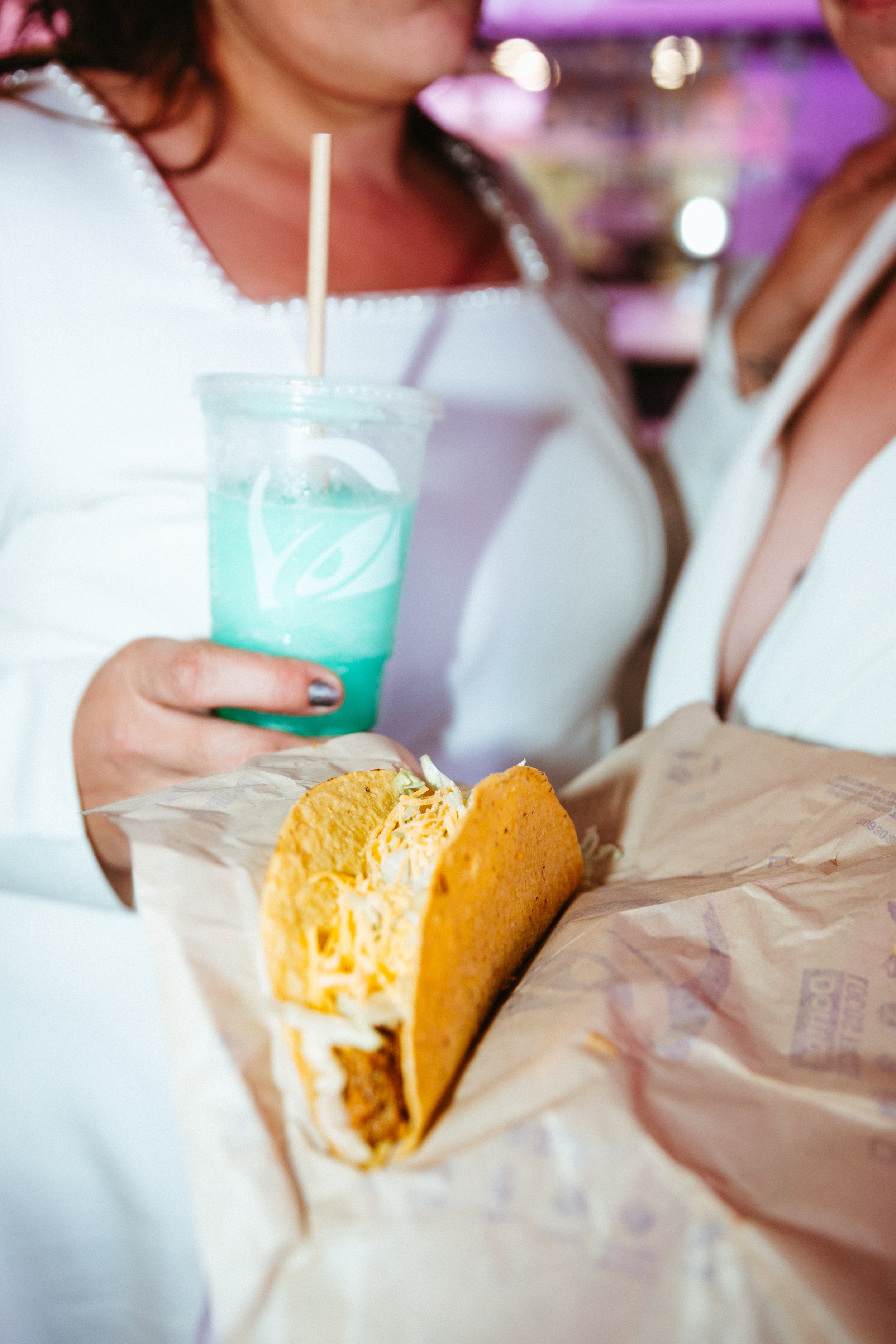 A person holding a taco with shredded cheese and meat, wrapped in paper, with a blue cocktail in the background.
