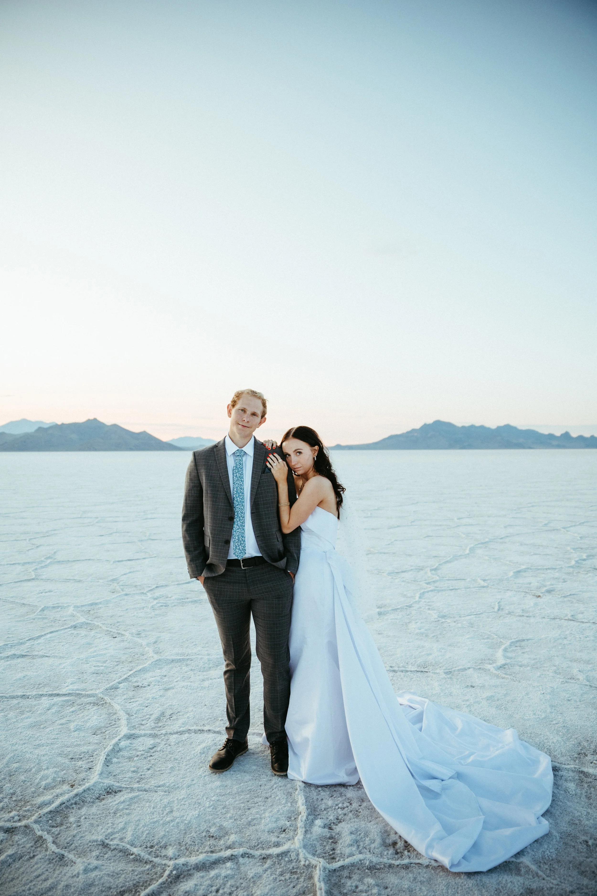 A couple dressed in wedding attire standing on a salt flat landscape with distant mountains in the background.