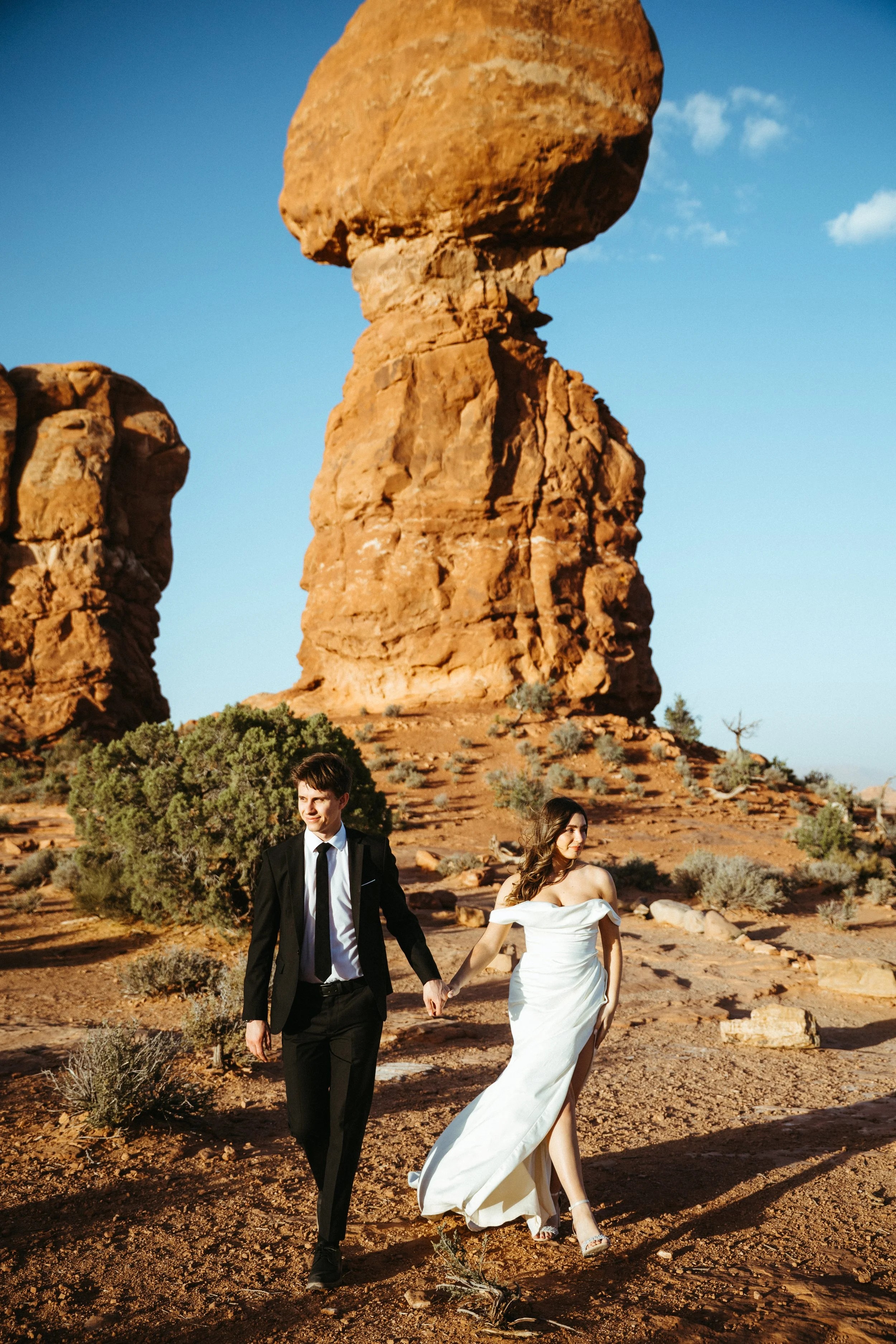A couple dressed in wedding attire holding hands while walking in a desert landscape with large rock formations and scattered shrubs under a clear blue sky.