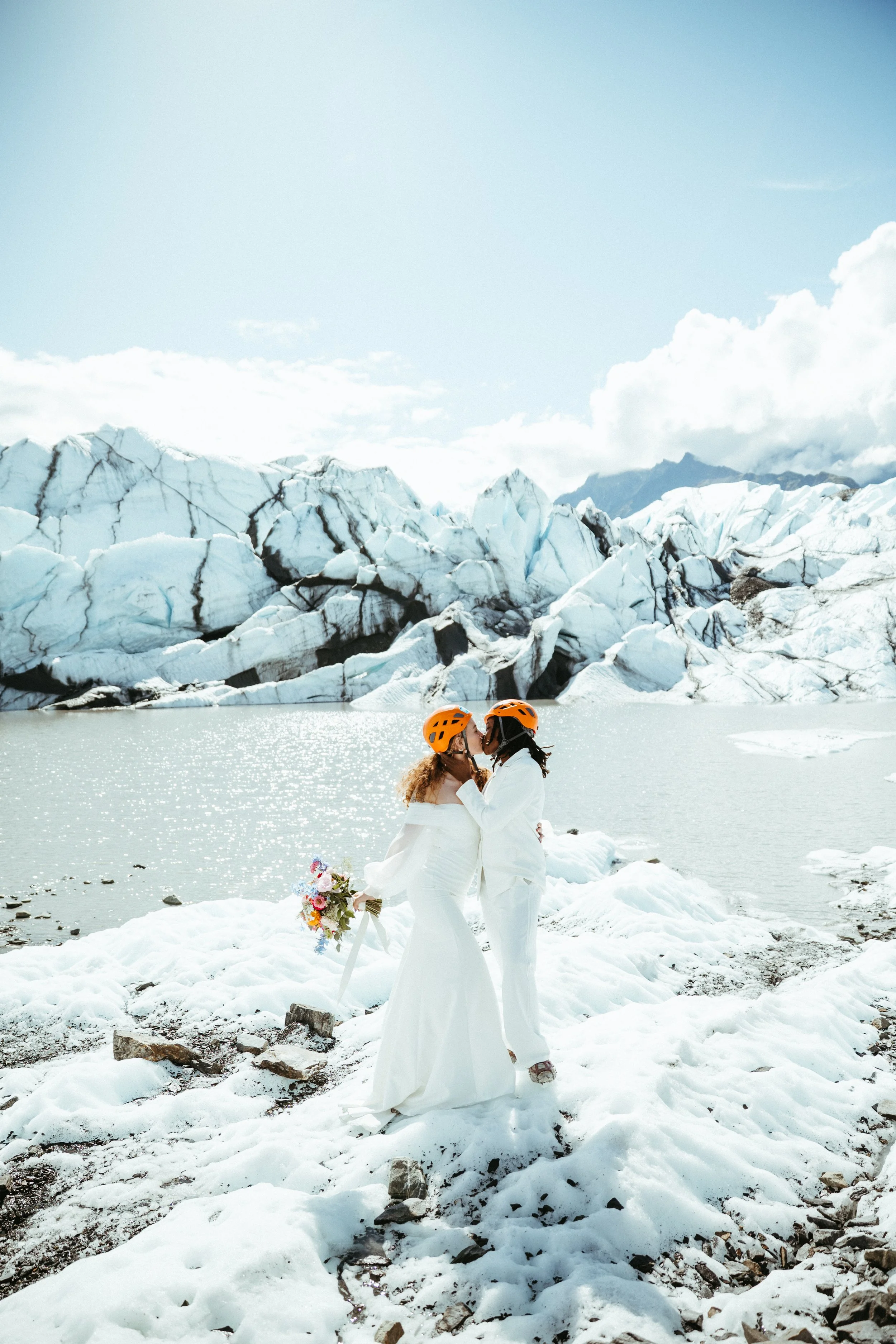 Two women in white wedding dresses and orange helmets sharing a kiss by a glacier lake, holding a bouquet, with icy mountains in the background.