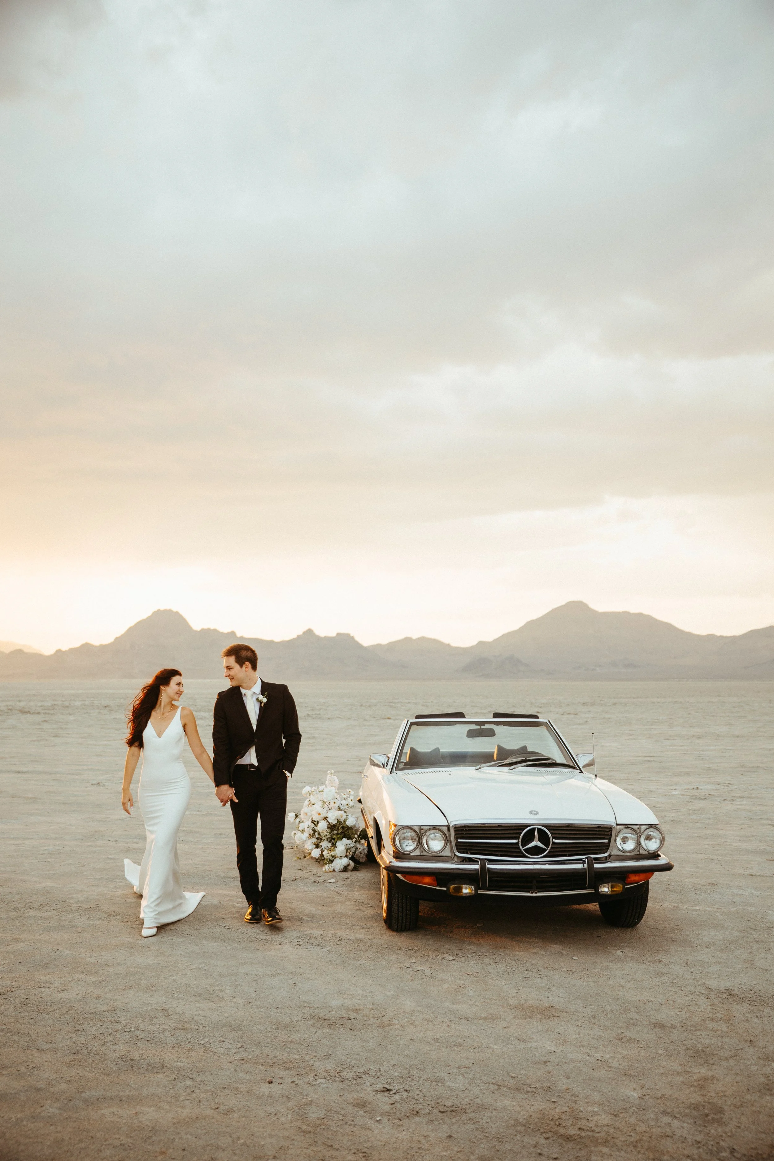 Bride and groom holding hands walking towards a vintage Mercedes-Benz convertible with flowers, on a barren landscape at sunset, mountains in the background.