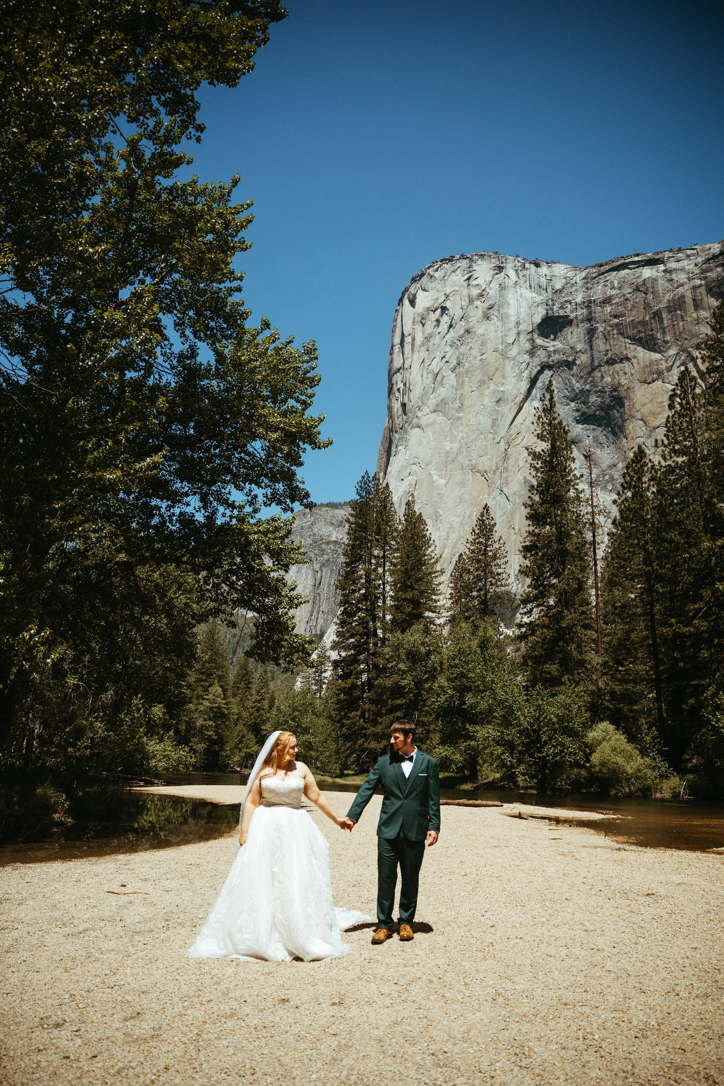 A bride and groom holding hands on a beach at a national park, with tall trees and a large granite cliff in the background.