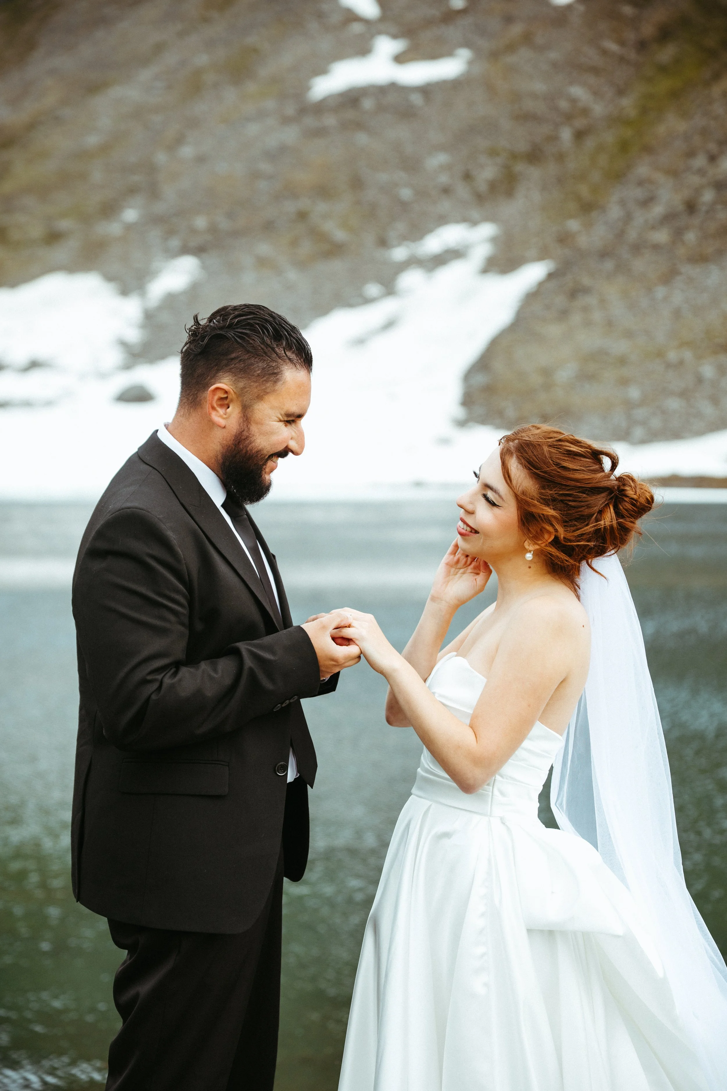 A bride and groom exchange vows outdoors by a mountain lake with snow patches and rocky slopes.