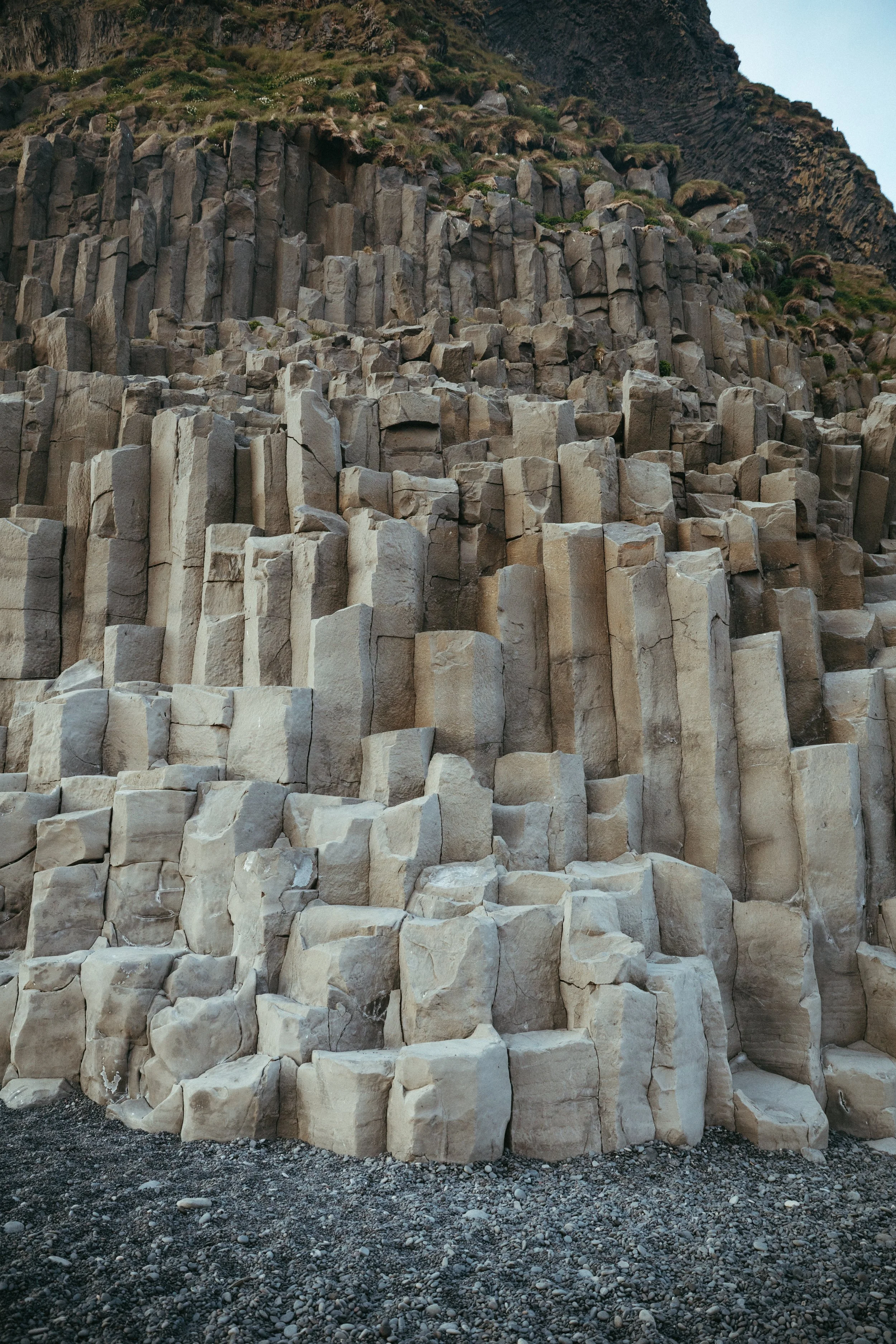 Columnar basalt rock formation with dark volcanic rocks at the top and lighter, hexagonal columns below, on a rocky ground.