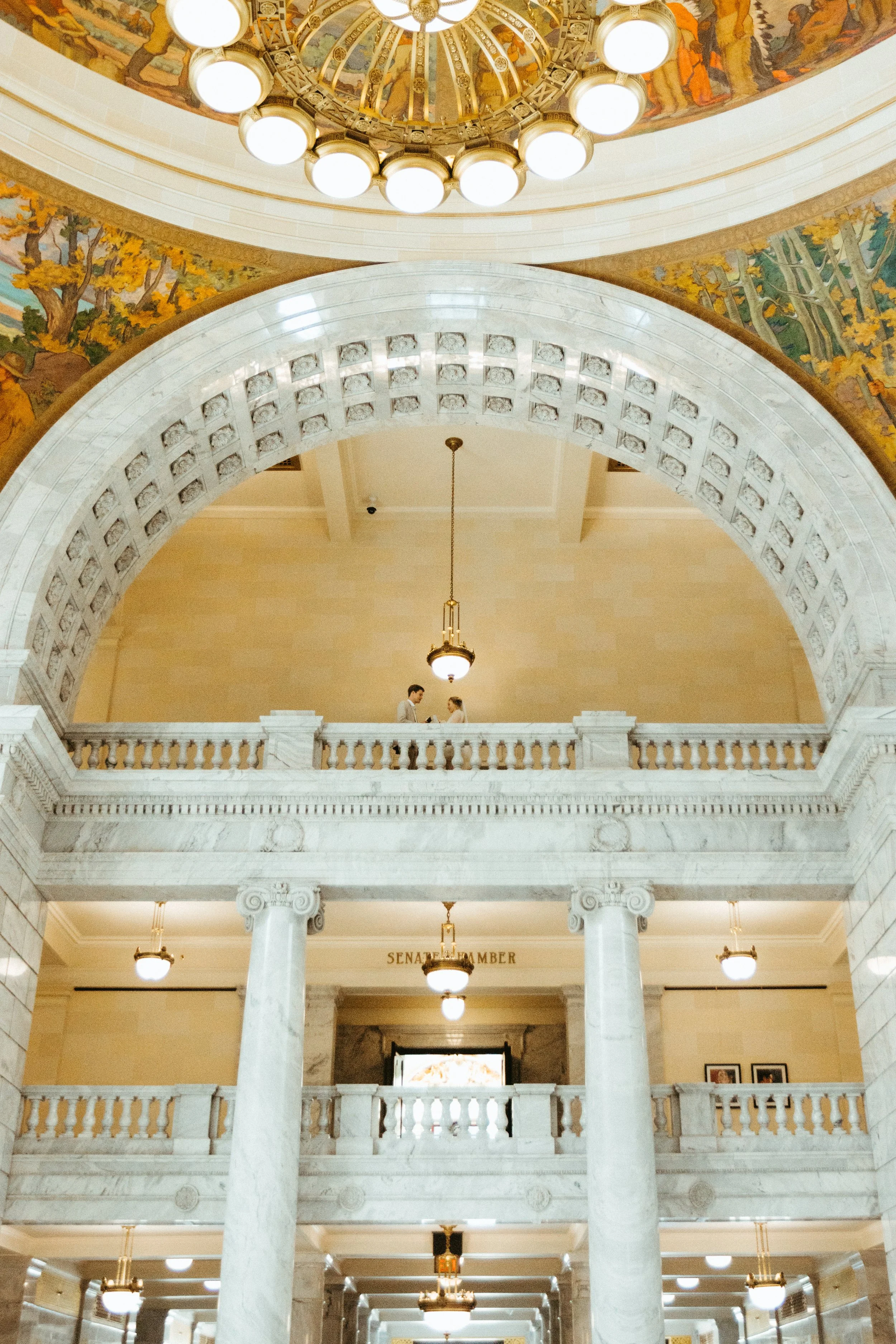 Interior of a government building with a marble staircase, arched balcony, ornate ceiling, and decorative lighting fixtures, with two people standing on the balcony.