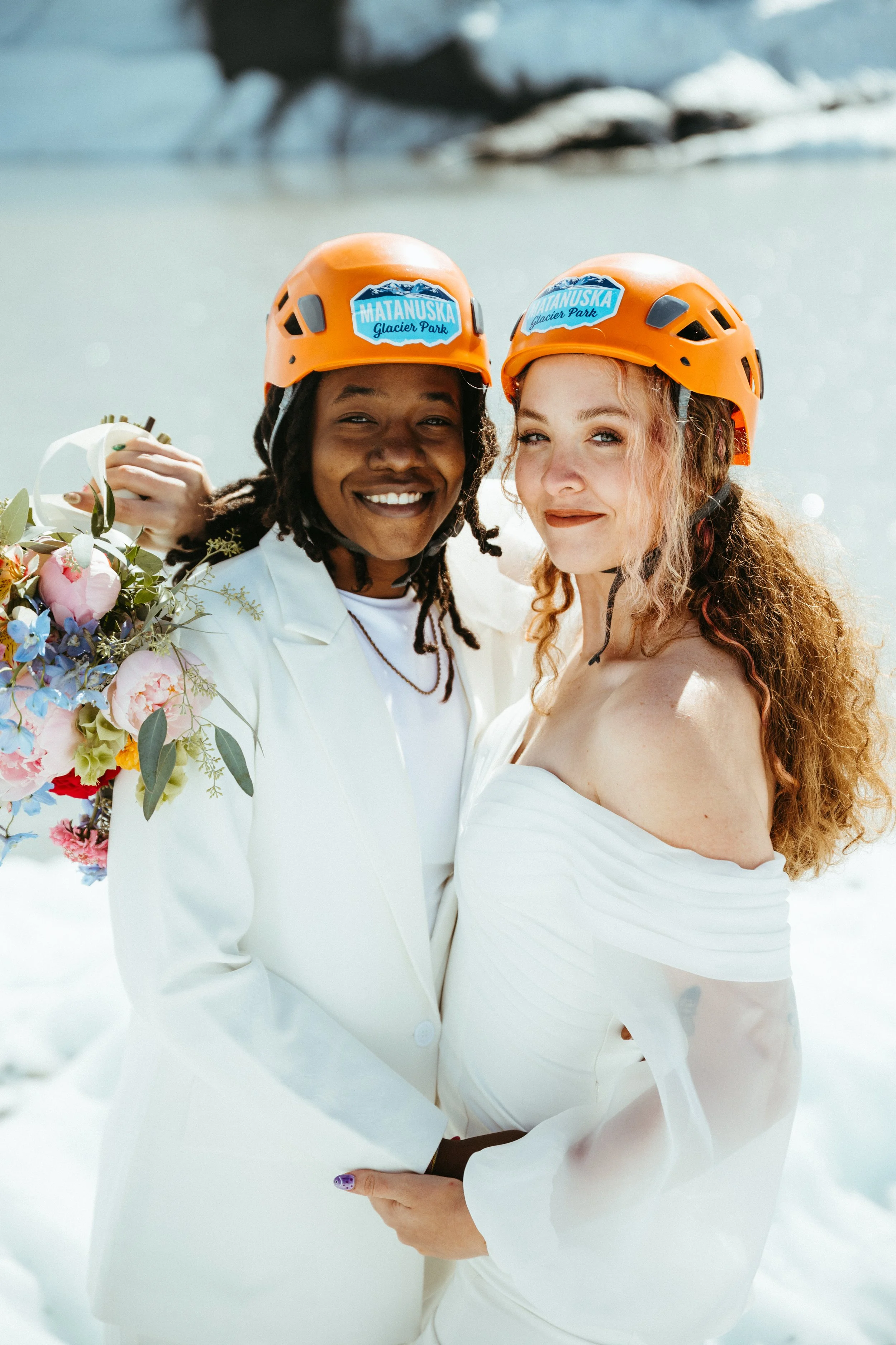 Two women in white wedding dresses holding each other, wearing orange helmets with 'Matanuska Glacier Park' labels, standing on snow with a glacier in the background.