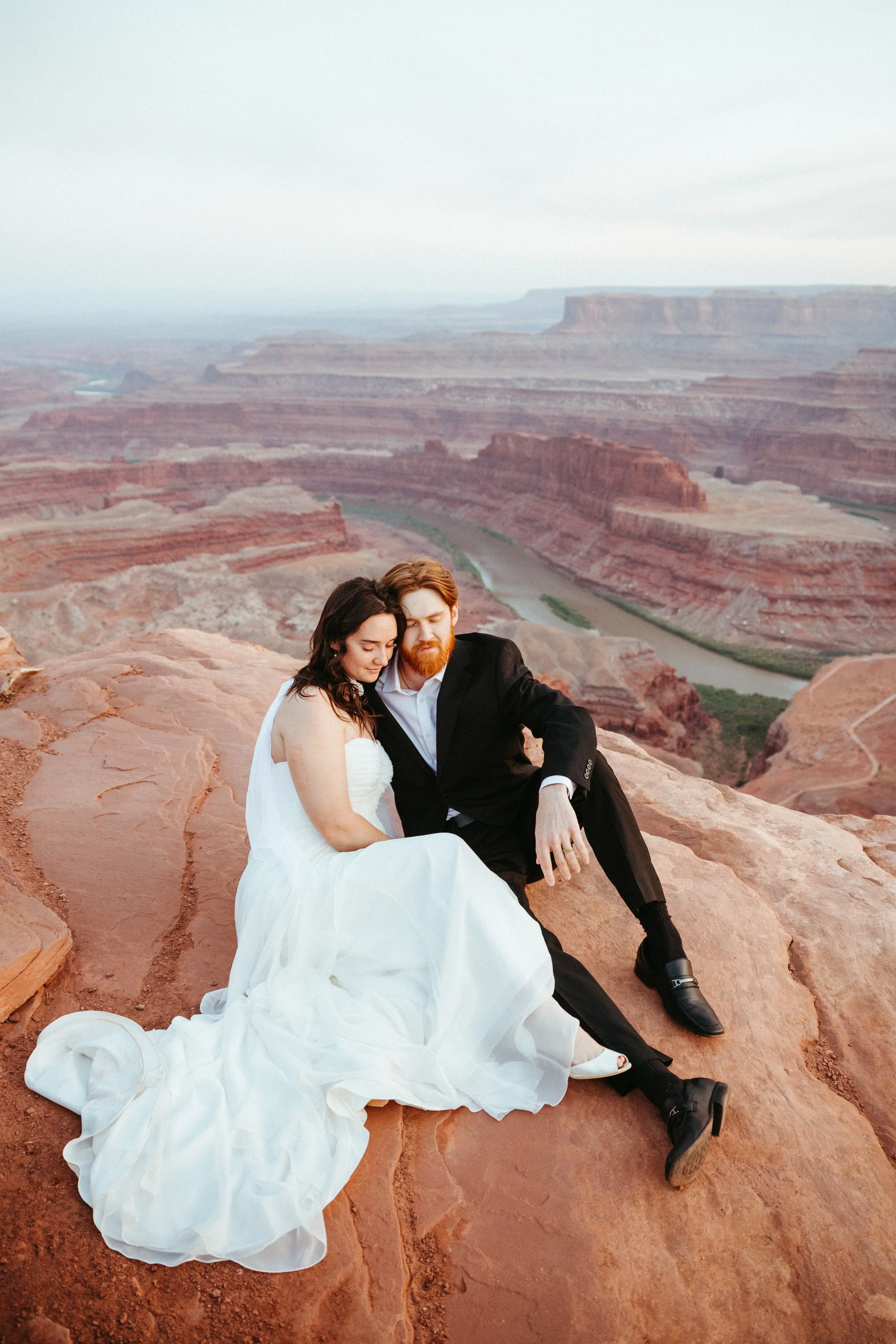 A bride in a white wedding dress and a groom in a black suit sitting on a rocky cliff with the Grand Canyon in the background.