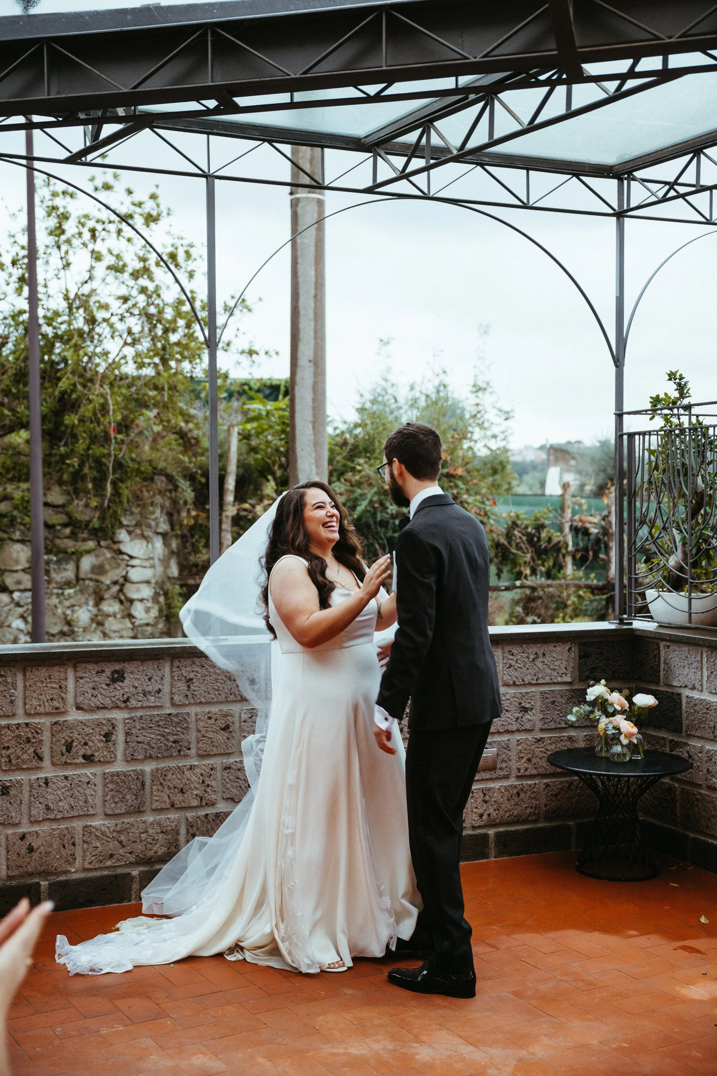 A bride in a white wedding dress and a groom in a black suit laughing and smiling during their wedding ceremony outdoors under a metal gazebo with greenery in the background.