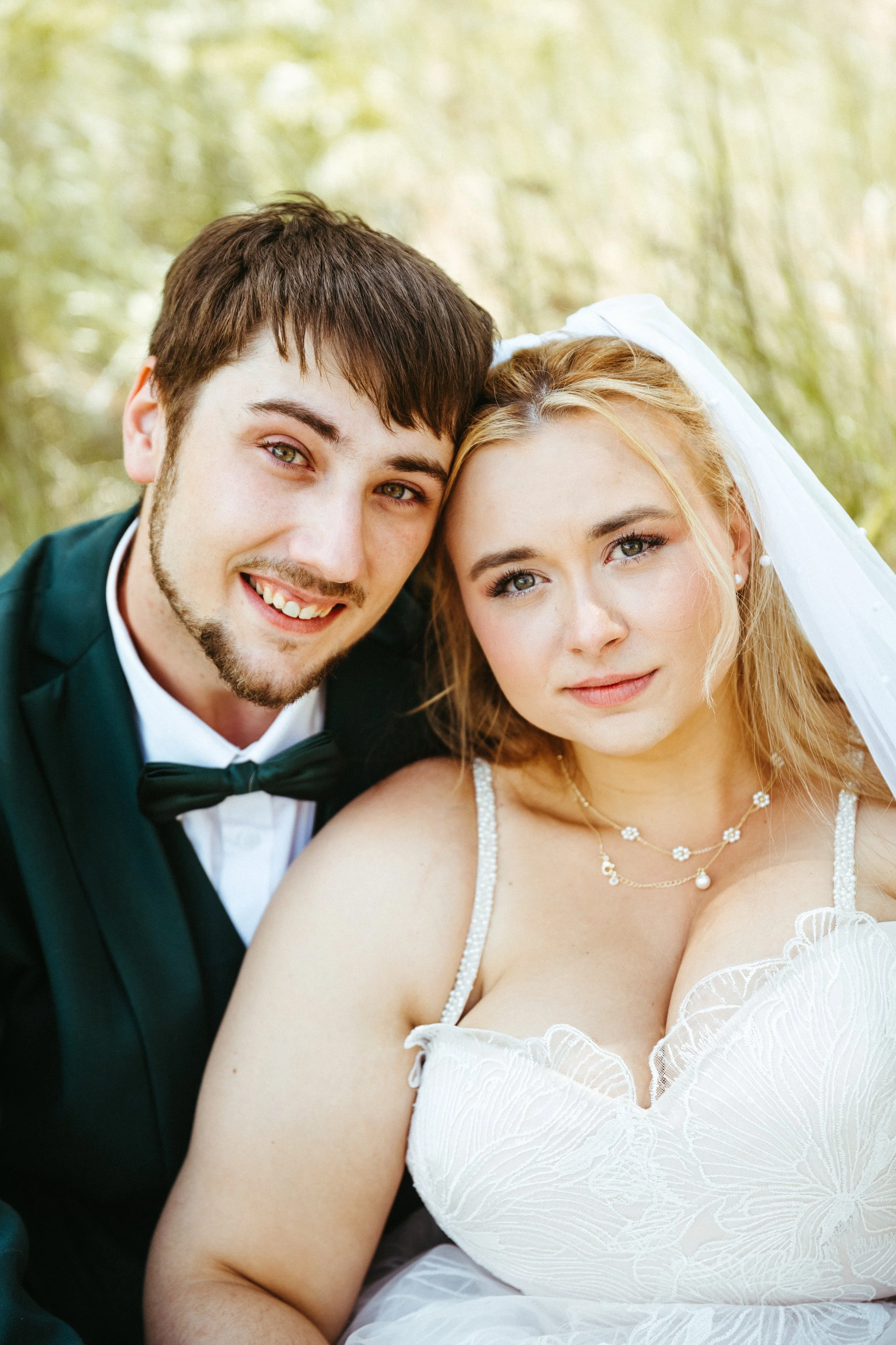 A newlywed couple, the groom in a tuxedo and the bride in a white wedding dress with jewelry, smiling and leaning close together outdoors with a blurred green background.