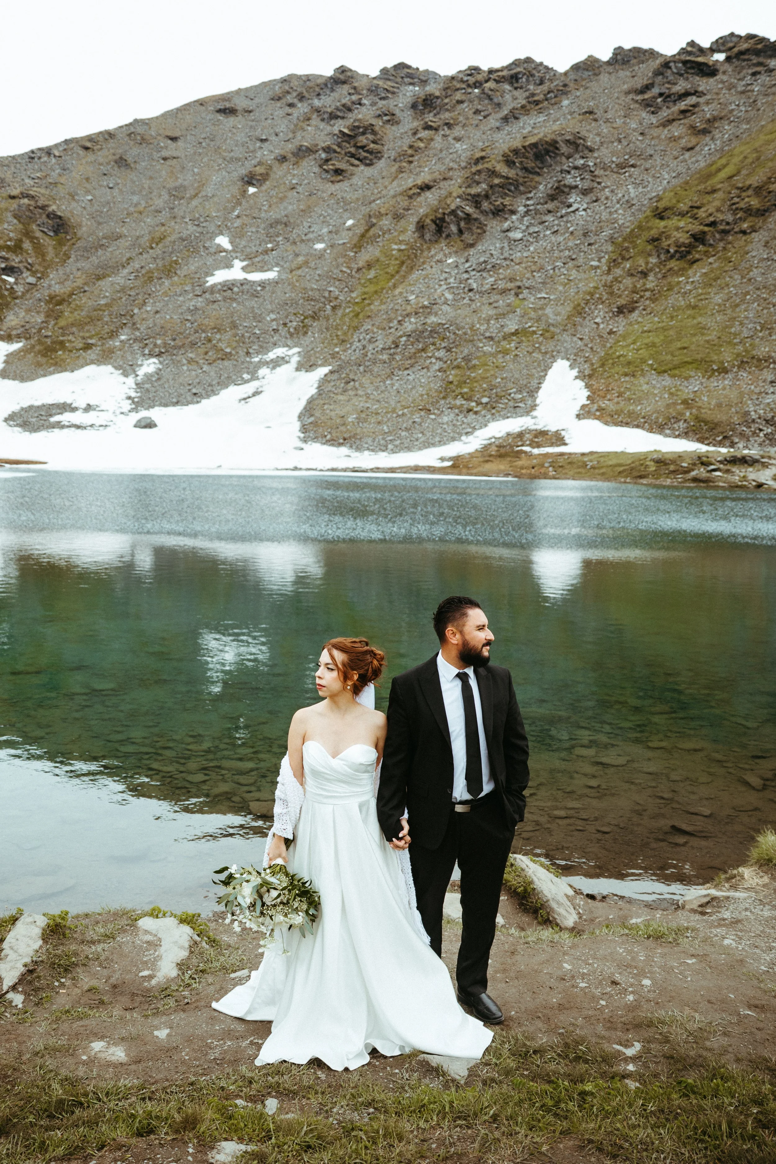 A bride and groom holding hands by a mountain lake, with snow-capped mountains in the background.