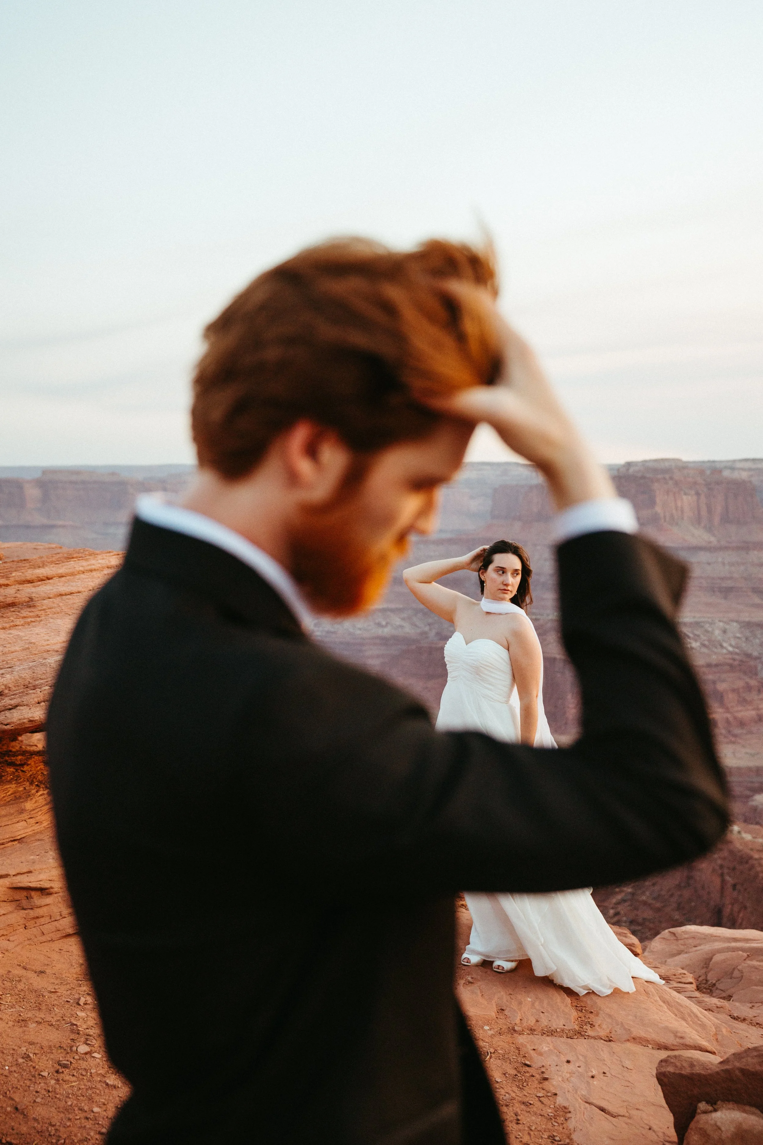 A man dressed in a black suit is in the foreground, with his hand in his hair, looking down. A woman in a white dress stands behind him at a scenic desert canyon, striking a pose with one hand on her head, looking at the camera.