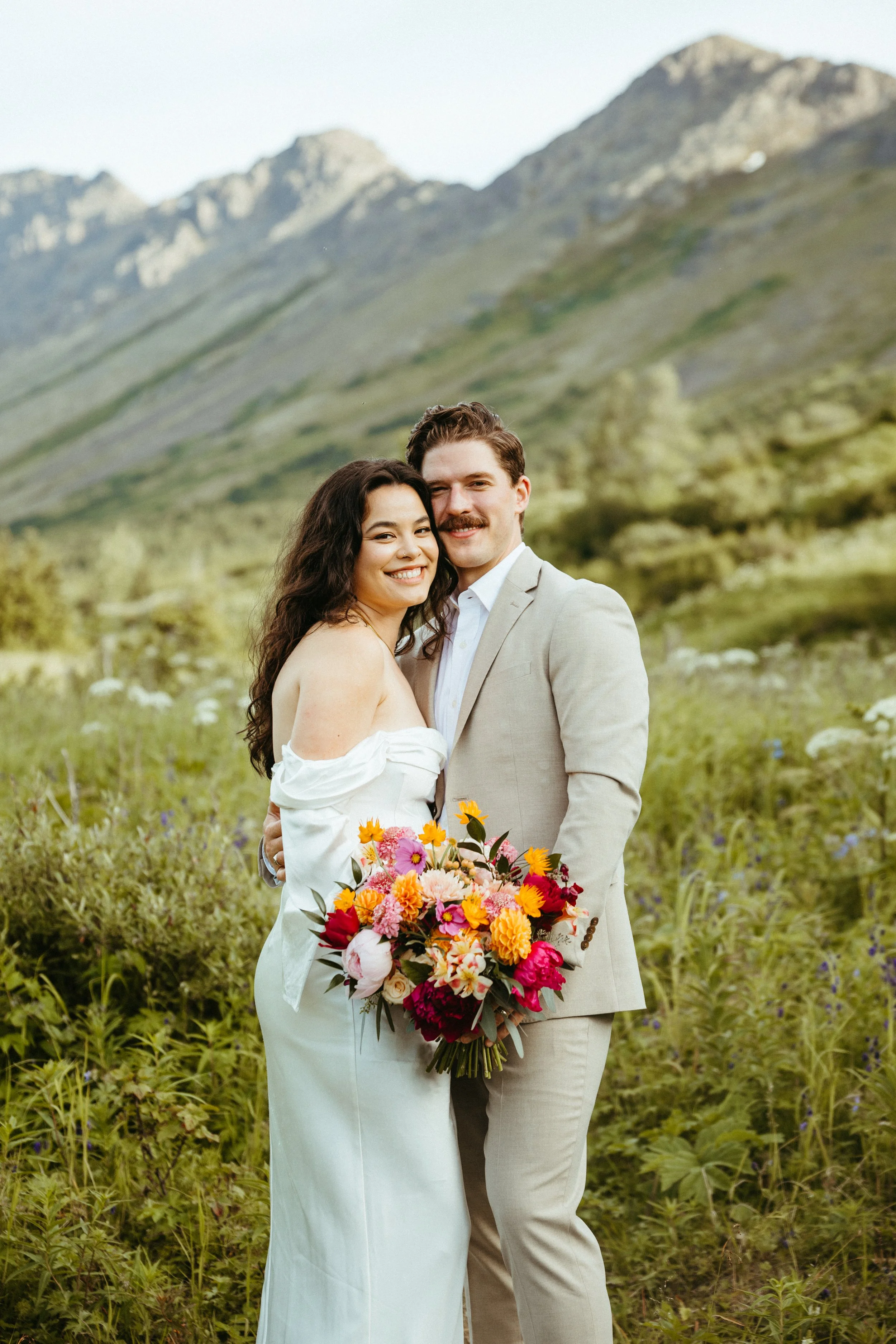 A happy couple dressed in wedding attire, standing outdoors in a lush, green field with mountains in the background, holding a colorful bouquet of flowers.