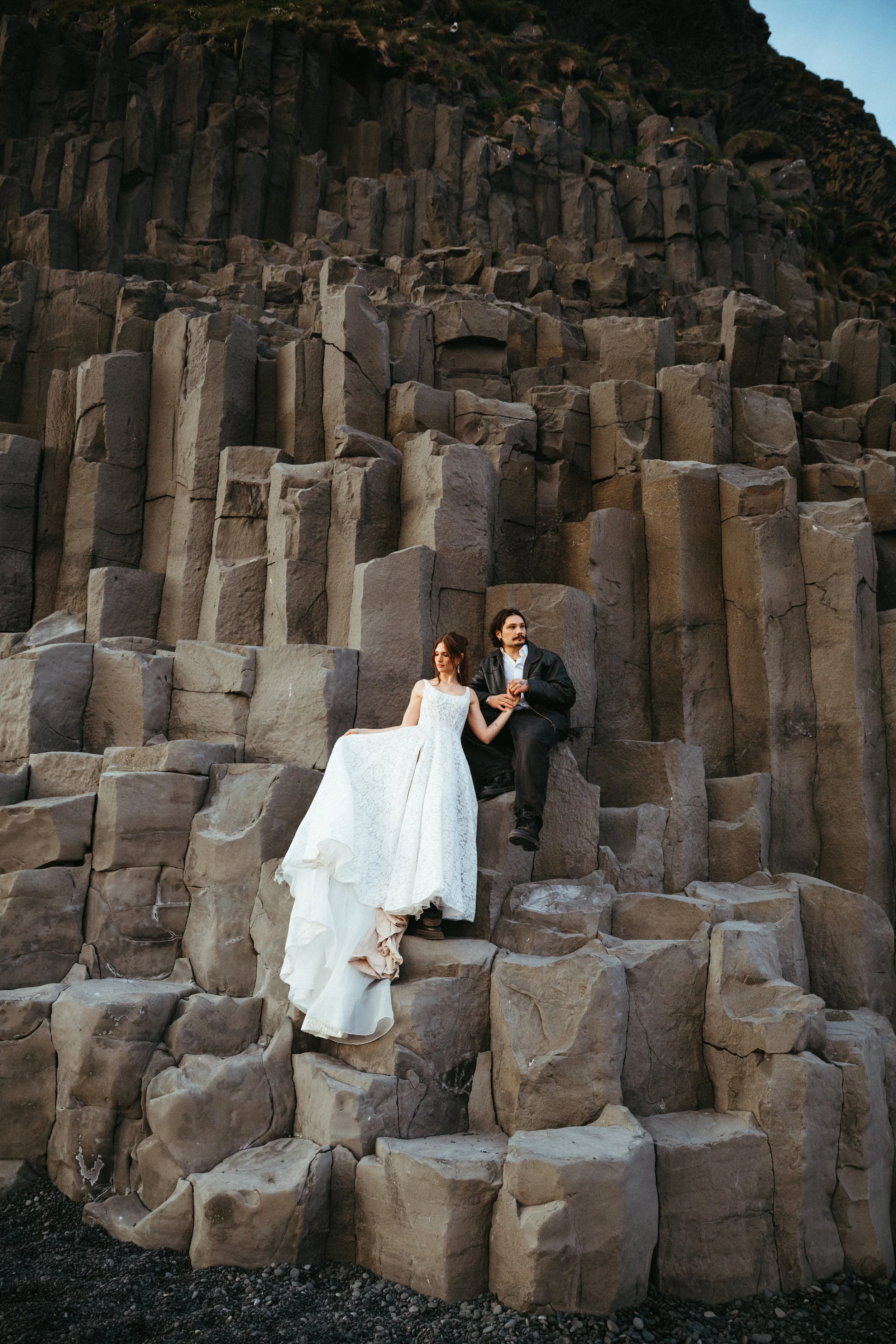 A man and a woman dressed in wedding attire pose on volcanic rocks with vertical columns. The woman wears a long white dress, and the man wears a black jacket and pants.