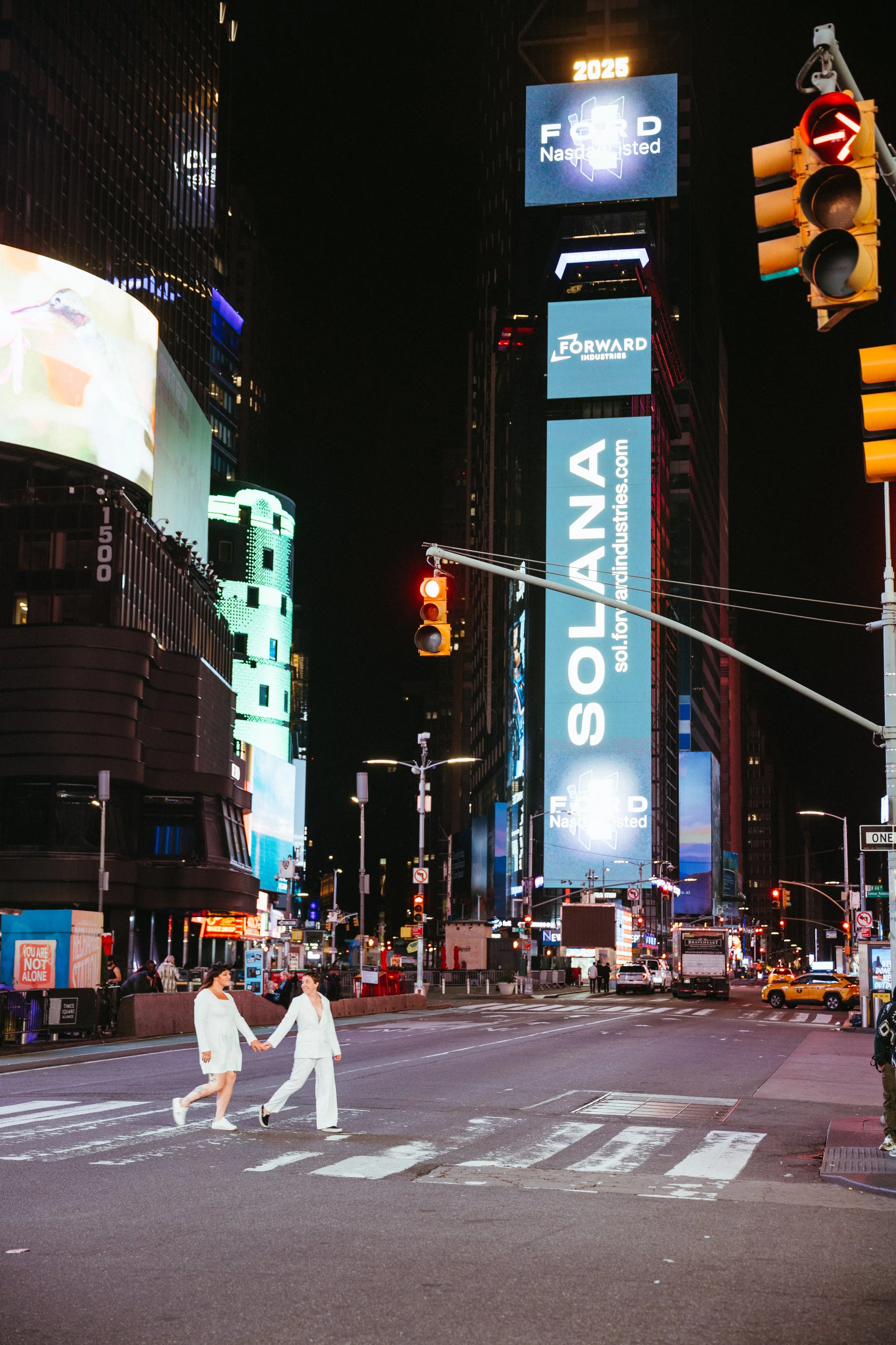Two women in white clothing holding hands and crossing the street at night in Times Square, New York City, with illuminated billboards and traffic lights.