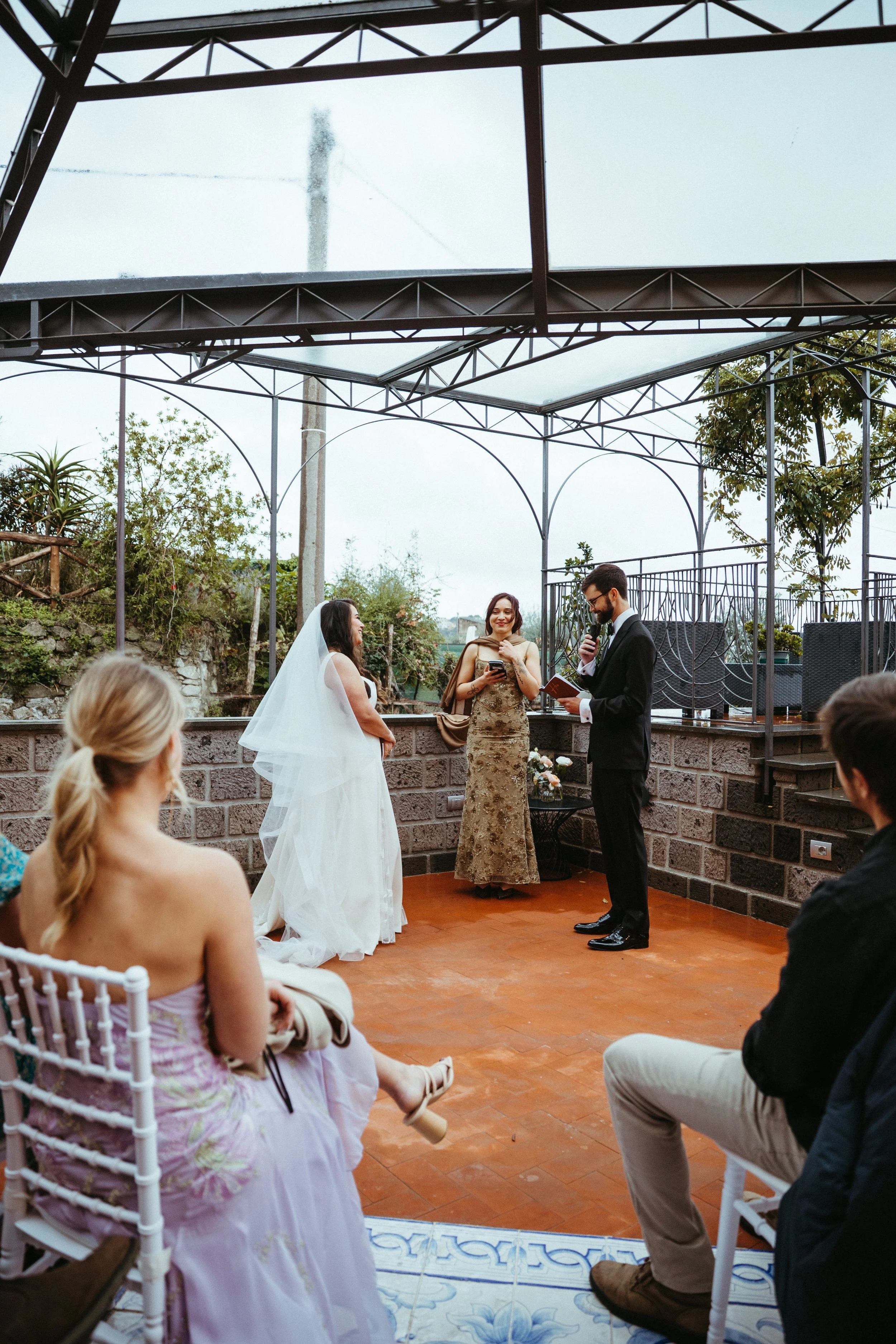 A wedding ceremony outdoors under a transparent roof with a bride in a white dress and veil, a groom in a black suit, and an officiant holding a microphone. Guests are seated nearby, watching the ceremony.