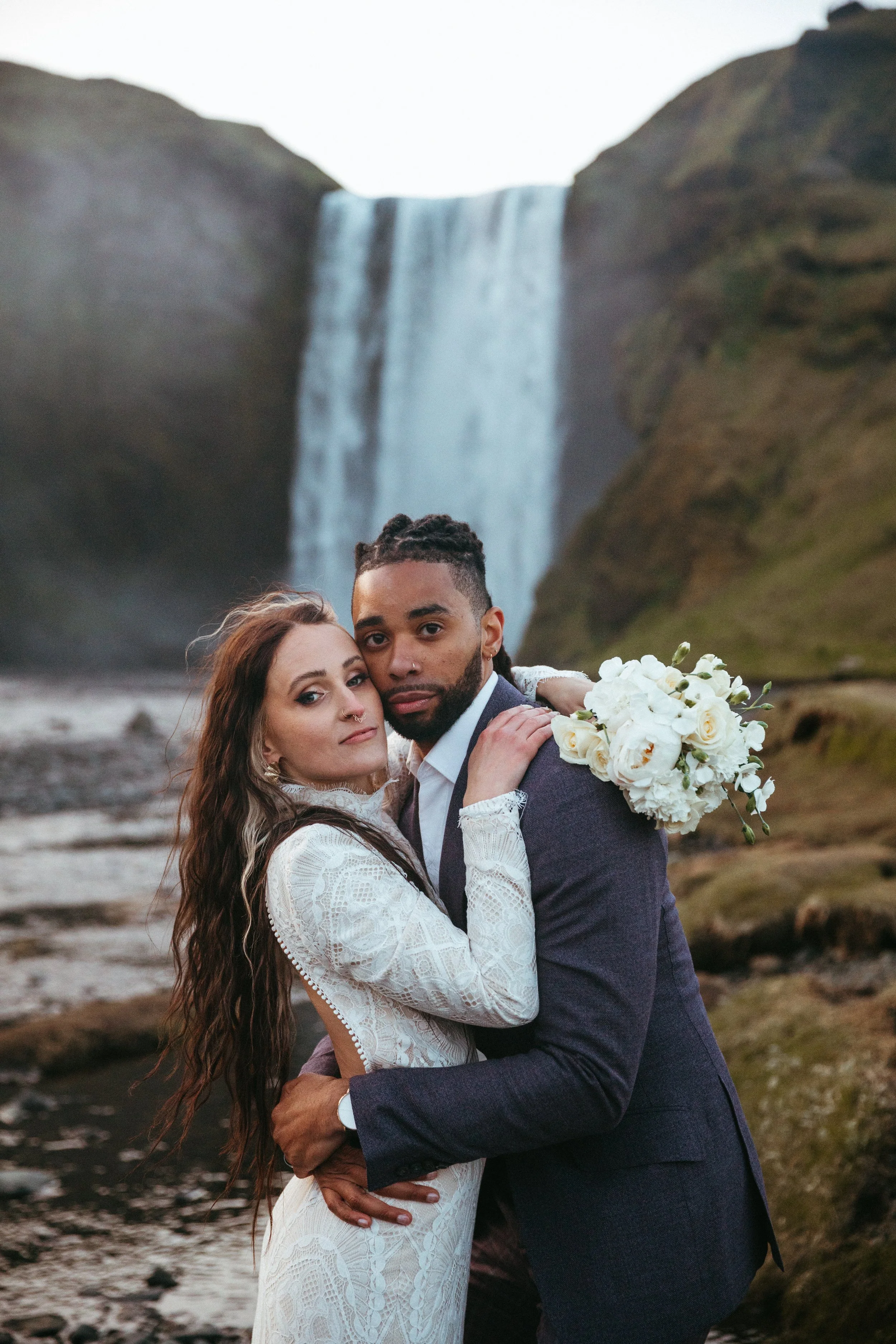A couple in wedding attire embracing outdoors with a large waterfall in the background.