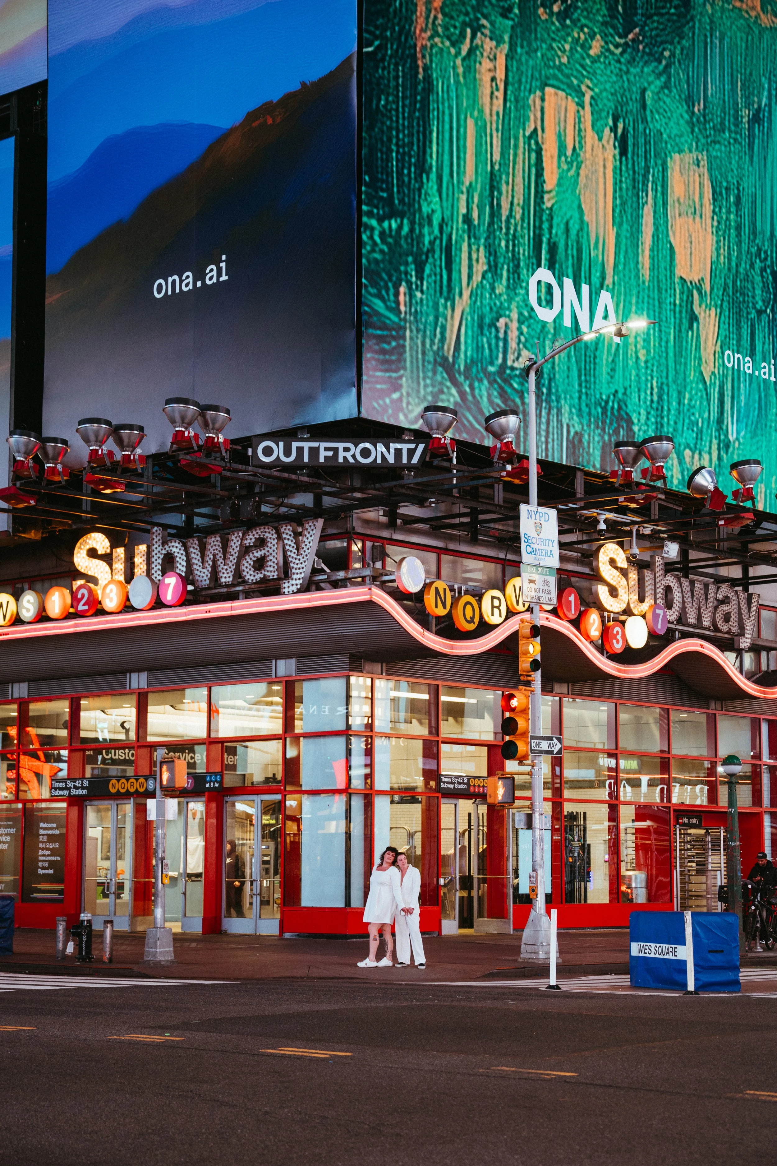Bright neon-lit entrance of New York City's Times Square subway station, with two women in white outfits standing at the crosswalk, surrounded by electronic billboards and urban signage.