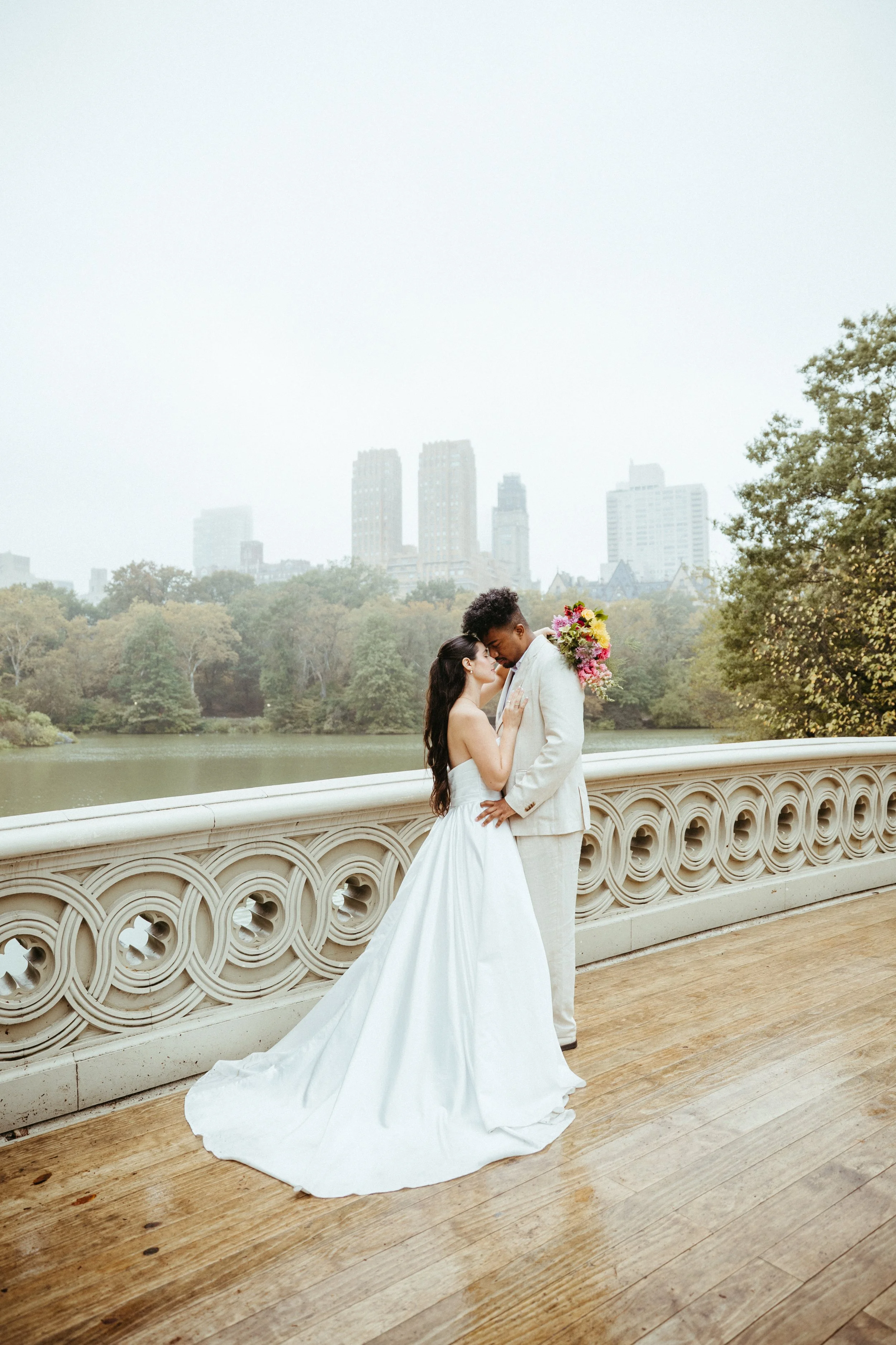 A newlywed couple sharing a kiss on a bridge overlooking a park with city skyline in the background.