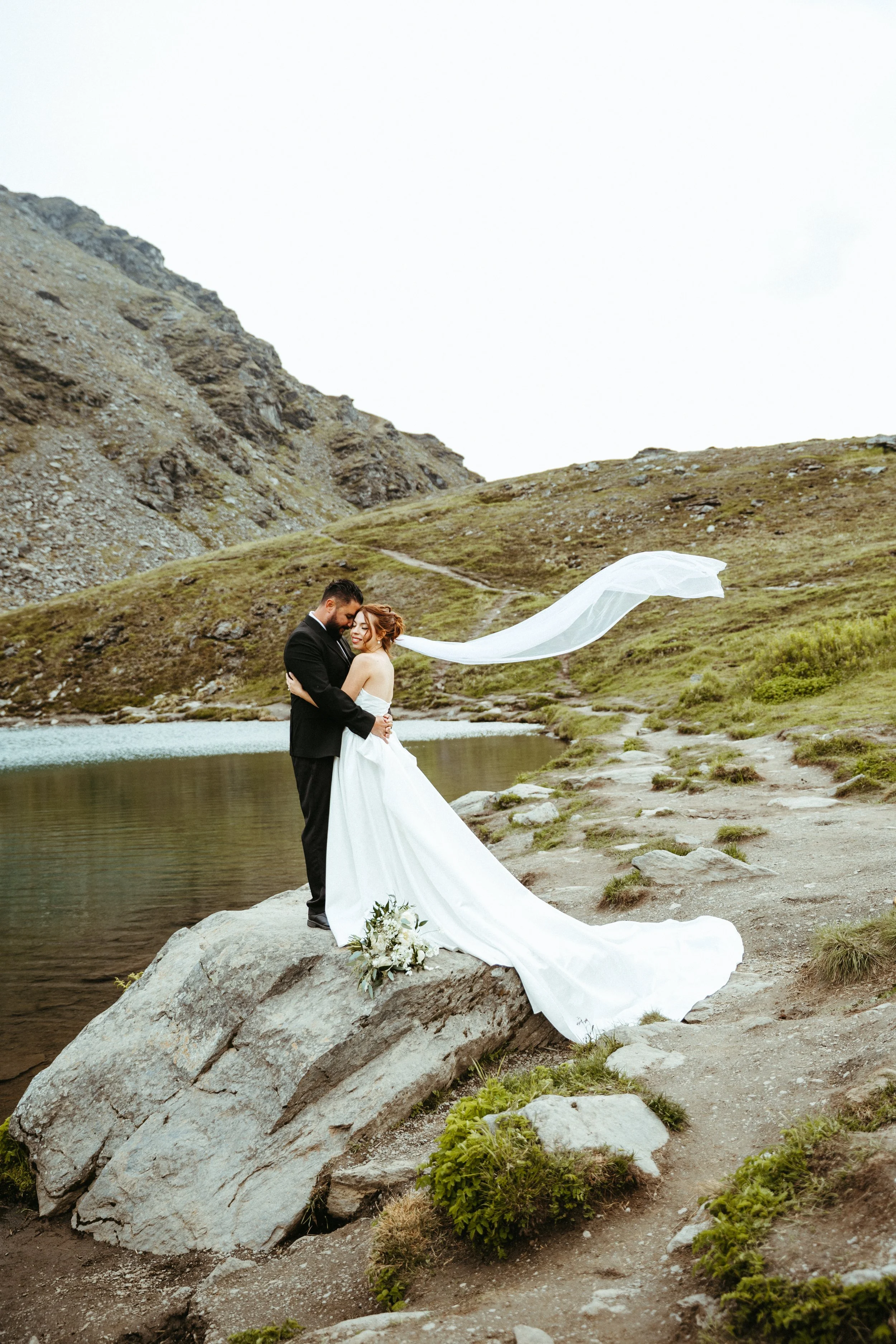 A bride and groom standing on a rock by a lake in a mountainous landscape, with the groom hugging the bride as she smiles, and her veil flowing in the wind.