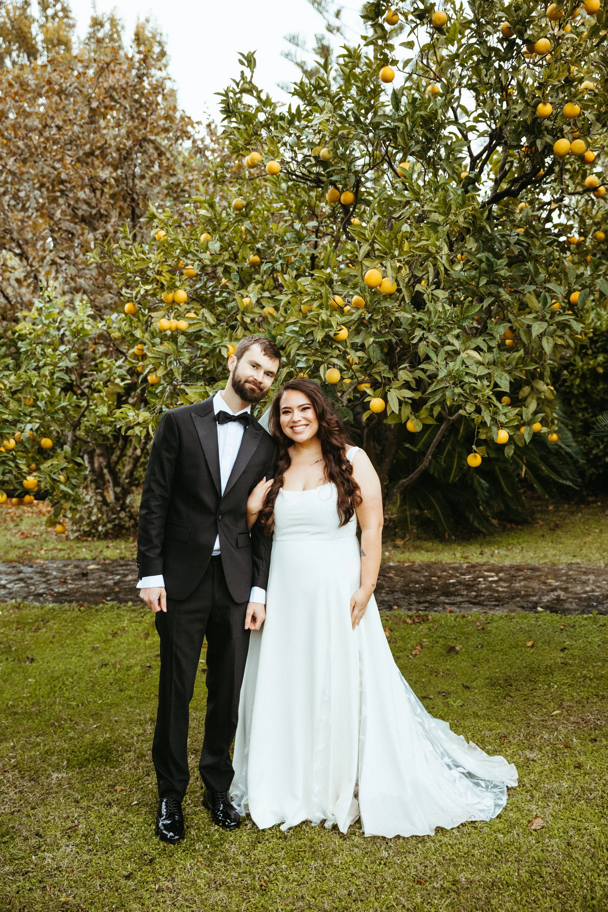 A wedding couple standing outside in front of an orange tree during daytime, with the groom in a black tuxedo and the bride in a white wedding gown.