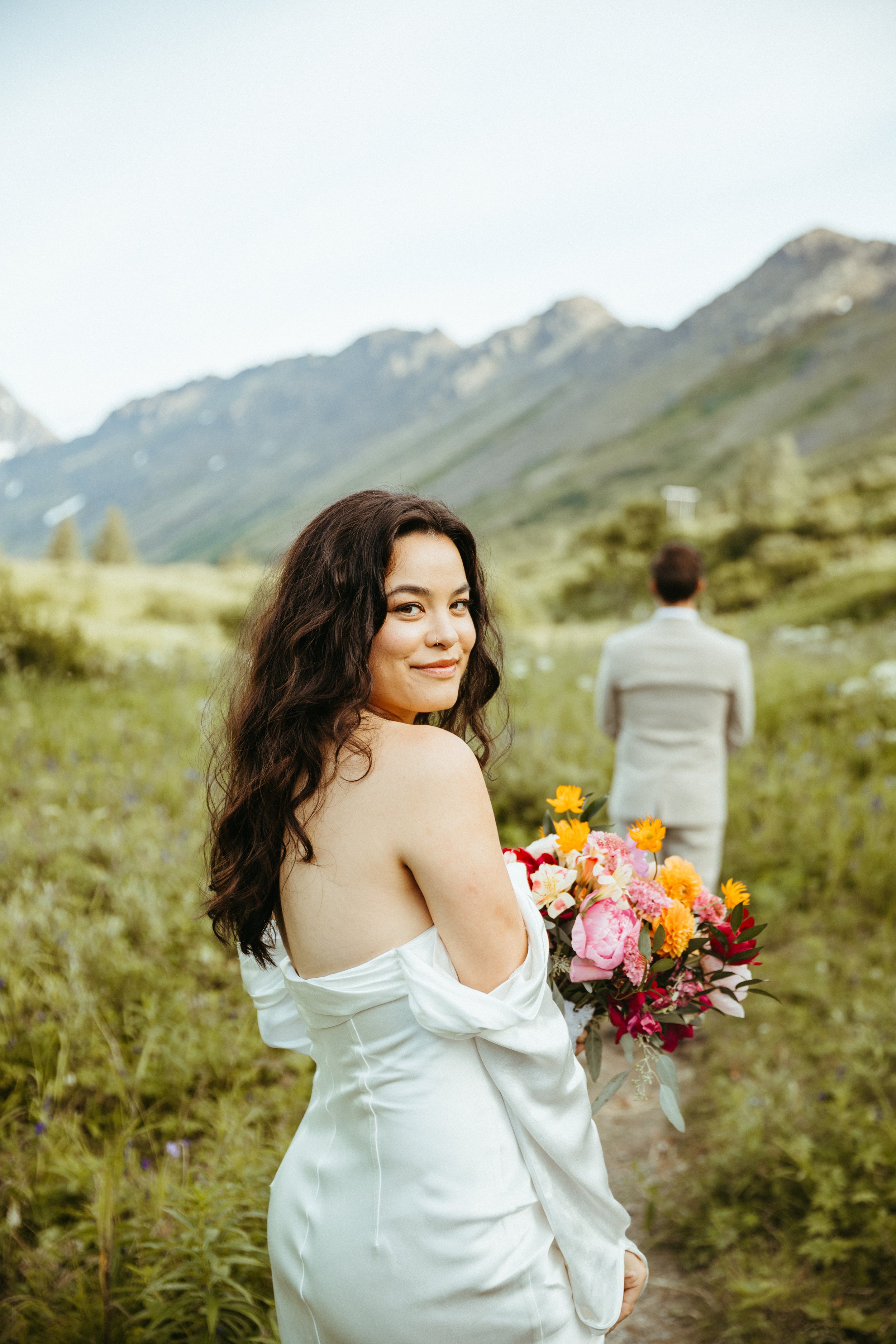 A woman in a white dress holding a bouquet of colorful flowers, standing outdoors in a green mountainous landscape, with a man in a gray suit in the background facing away.