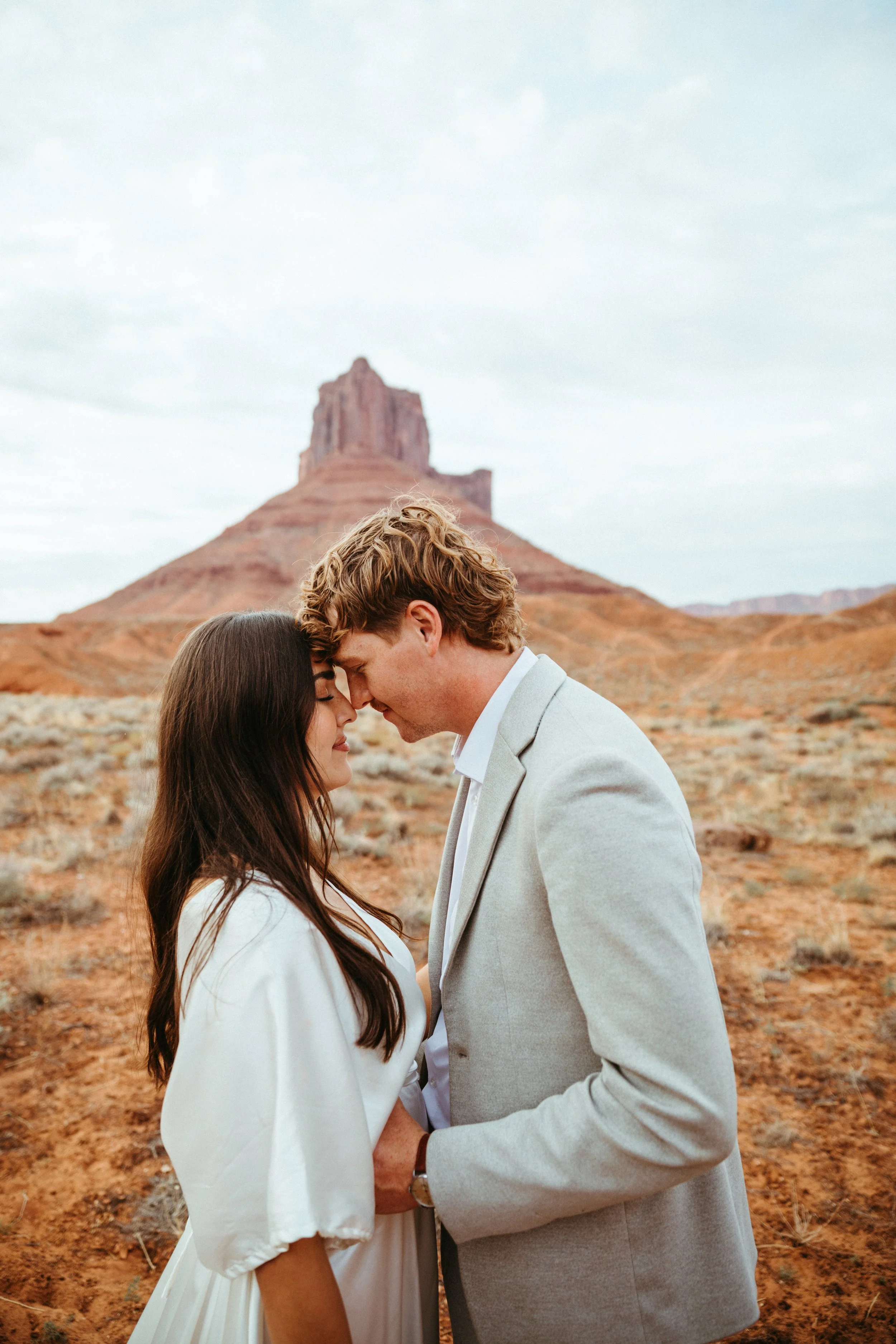 A couple faces each other with foreheads touching in a desert landscape with a large rock formation in the background.
