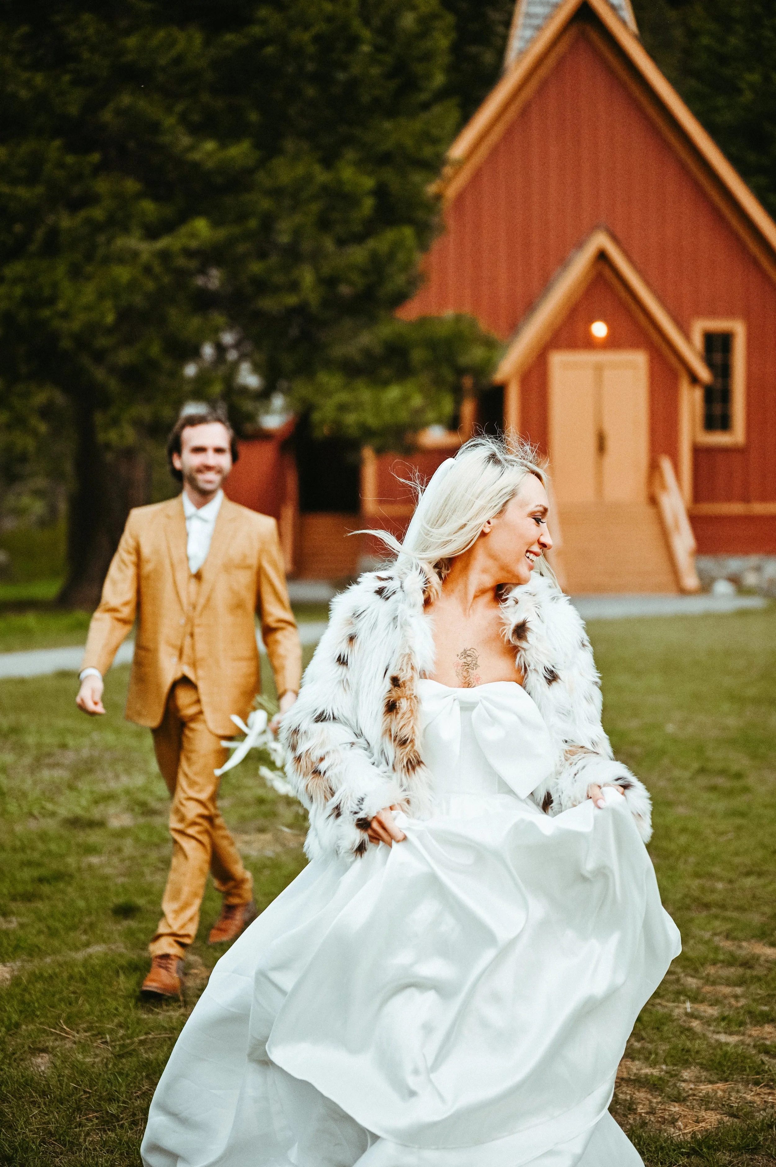 A woman in a white wedding dress and a fur coat running ahead of a man in a suit, with a small red wooden church in the background.
