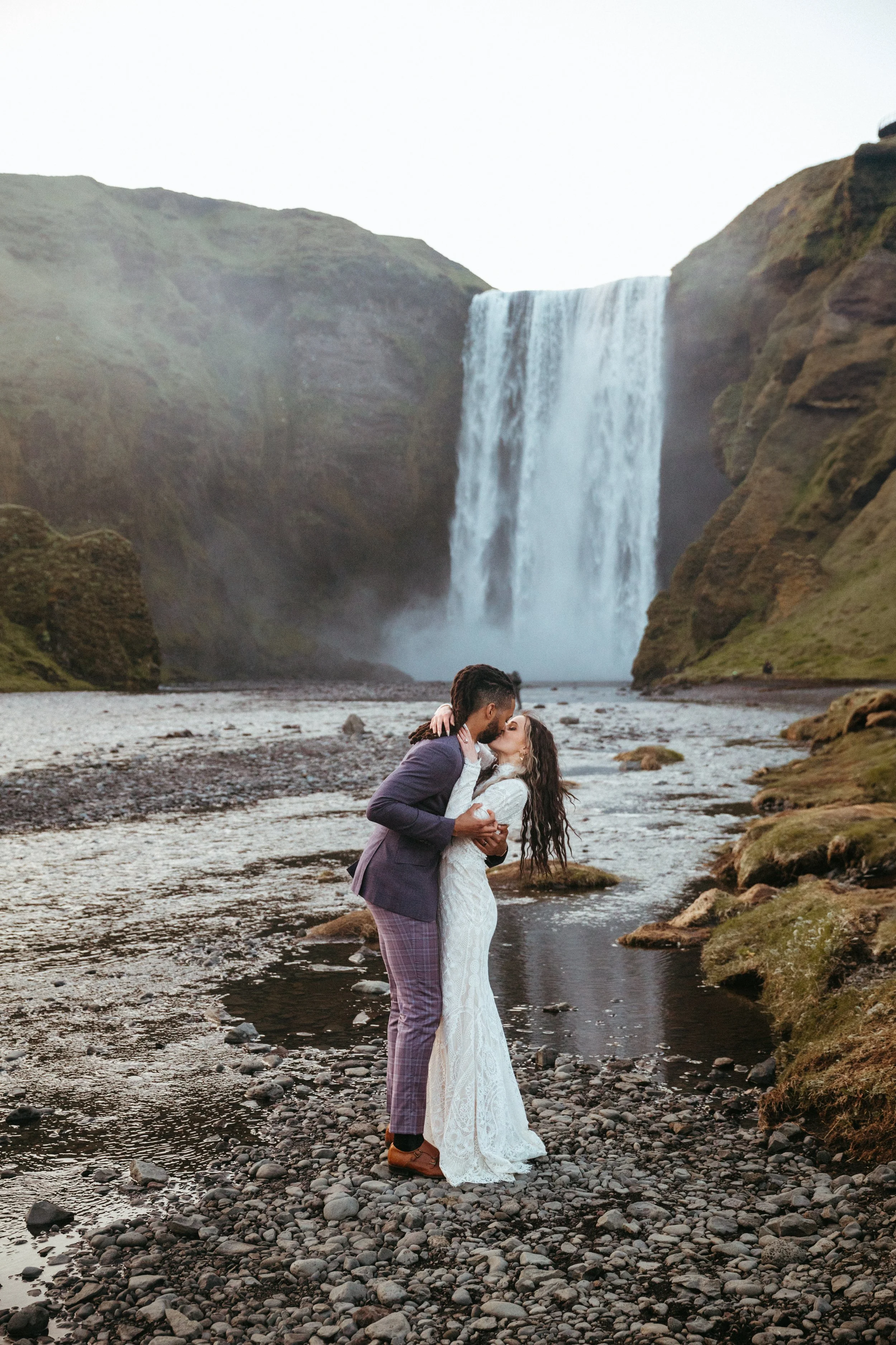 A couple in wedding attire sharing a kiss on rocky ground near a waterfall.