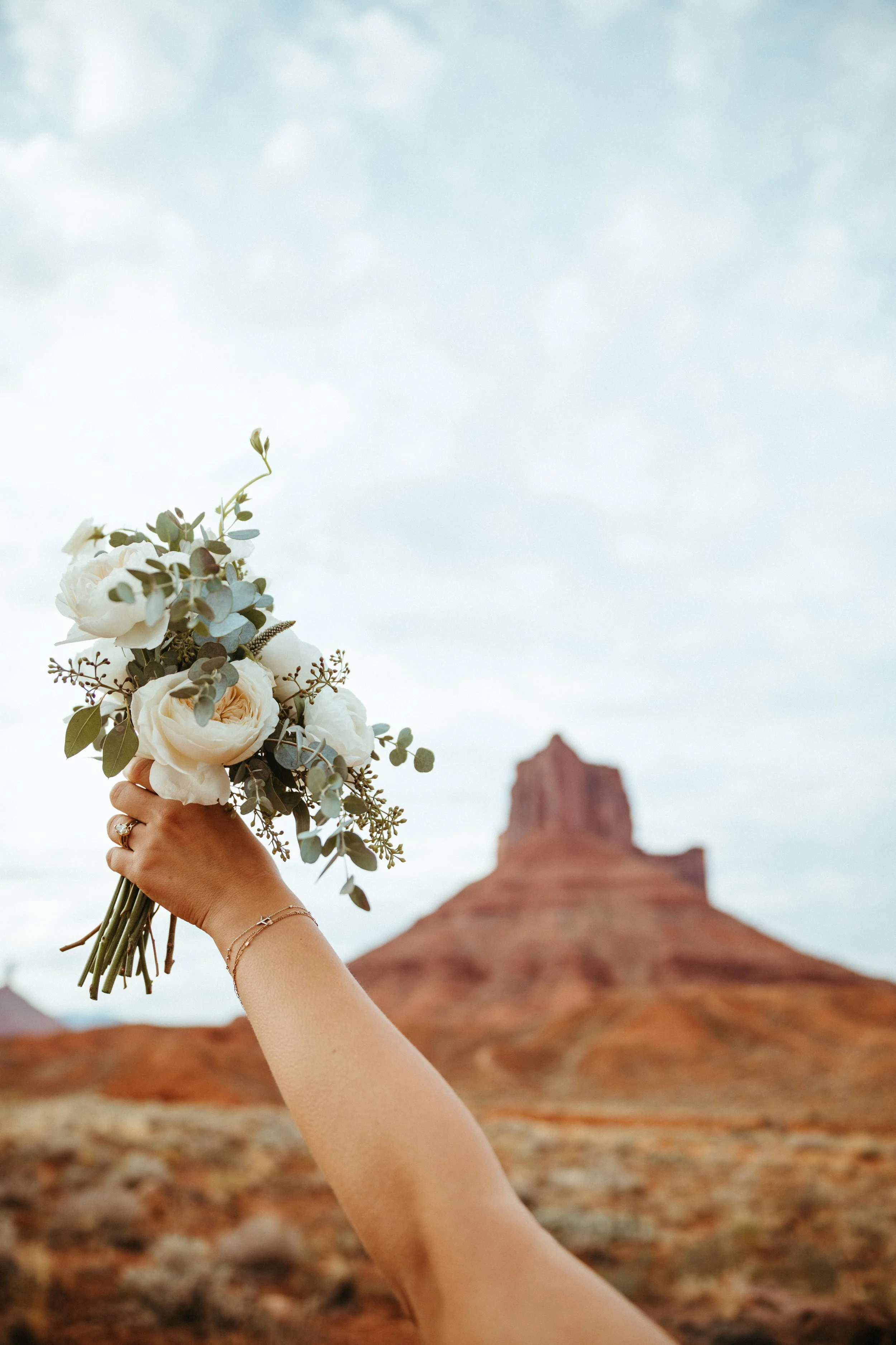 A hand holding a bouquet of white flowers with greenery, with a desert landscape and a prominent red rock formation in the background.