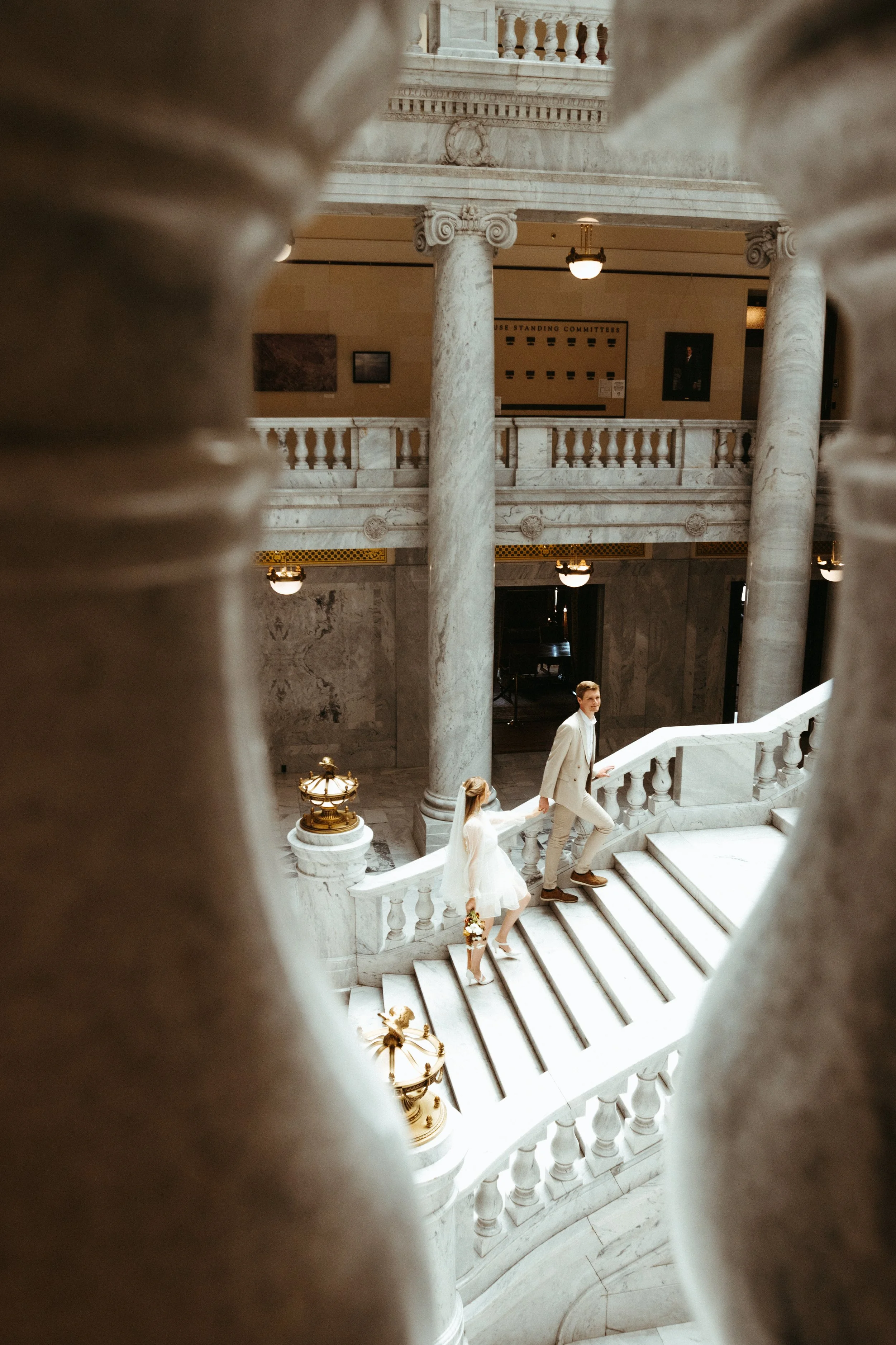 A bride and groom walking up the marble stairs inside a grand, historic building with large columns and ornate details.