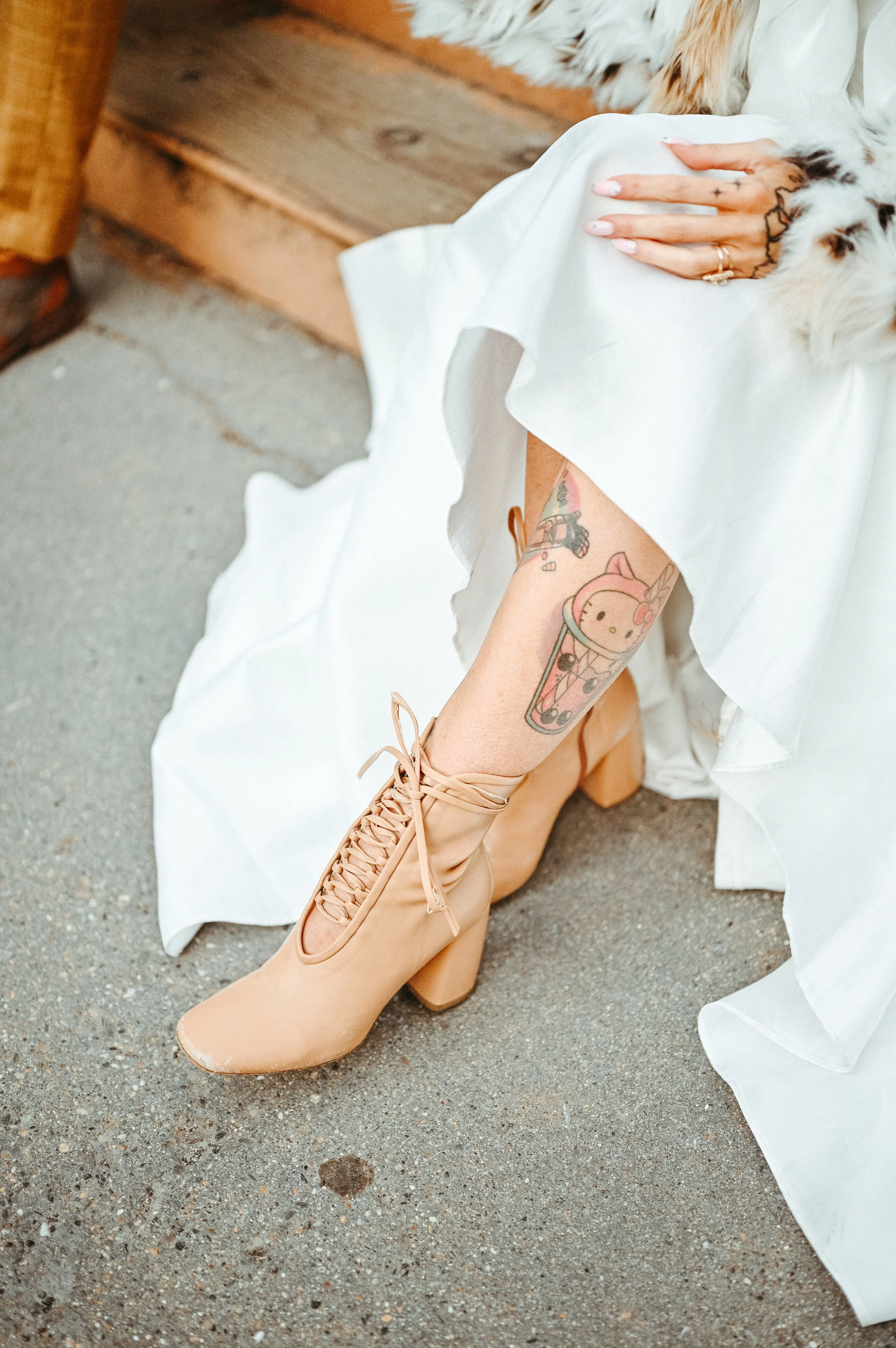 A person with tattooed legs wearing beige lace-up high-heeled boots, sitting on a concrete surface near wooden steps, partially covered by a white dress.