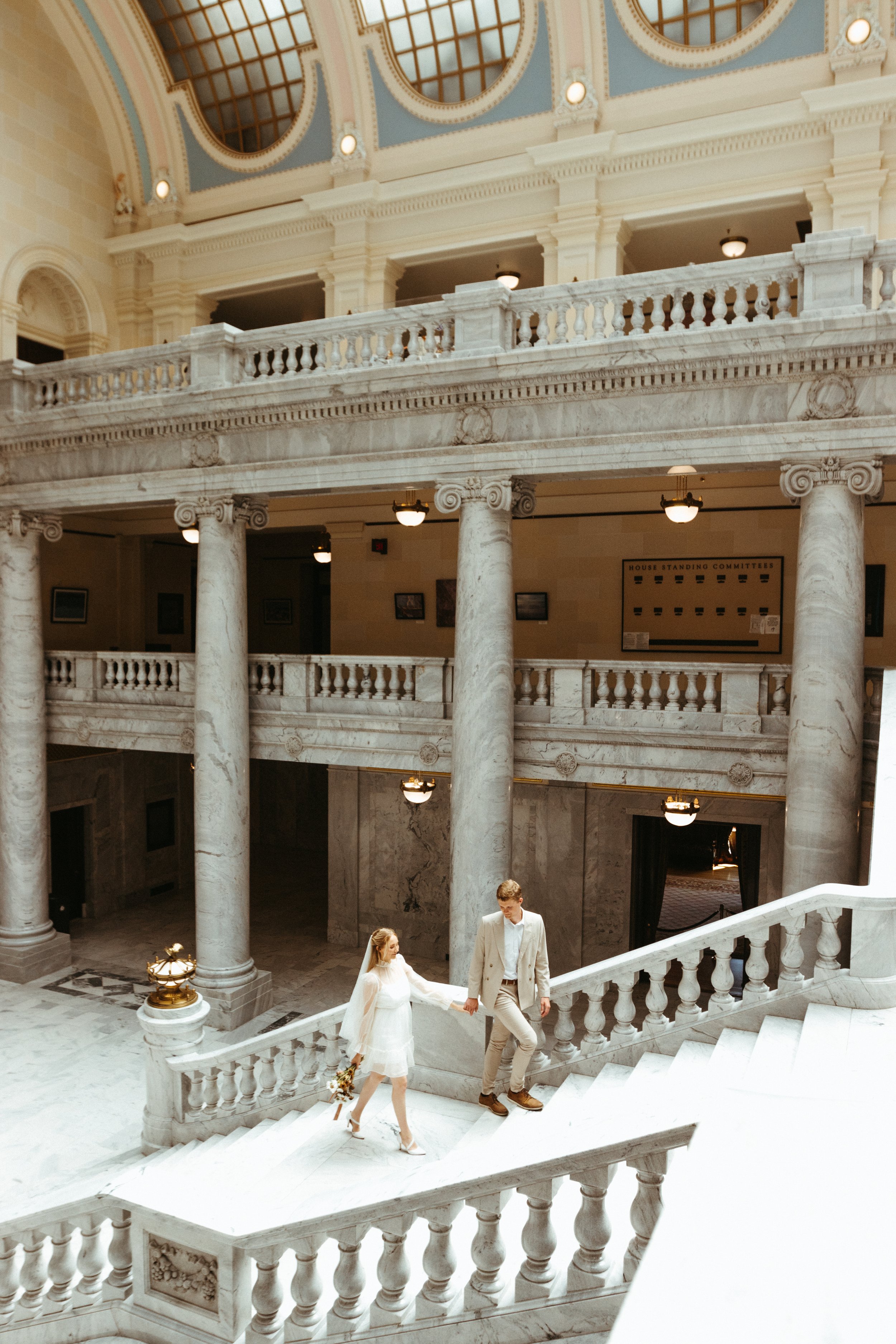A bride and groom holding hands and walking down a marble staircase inside a grand, historic building with columns and ornate architectural details.