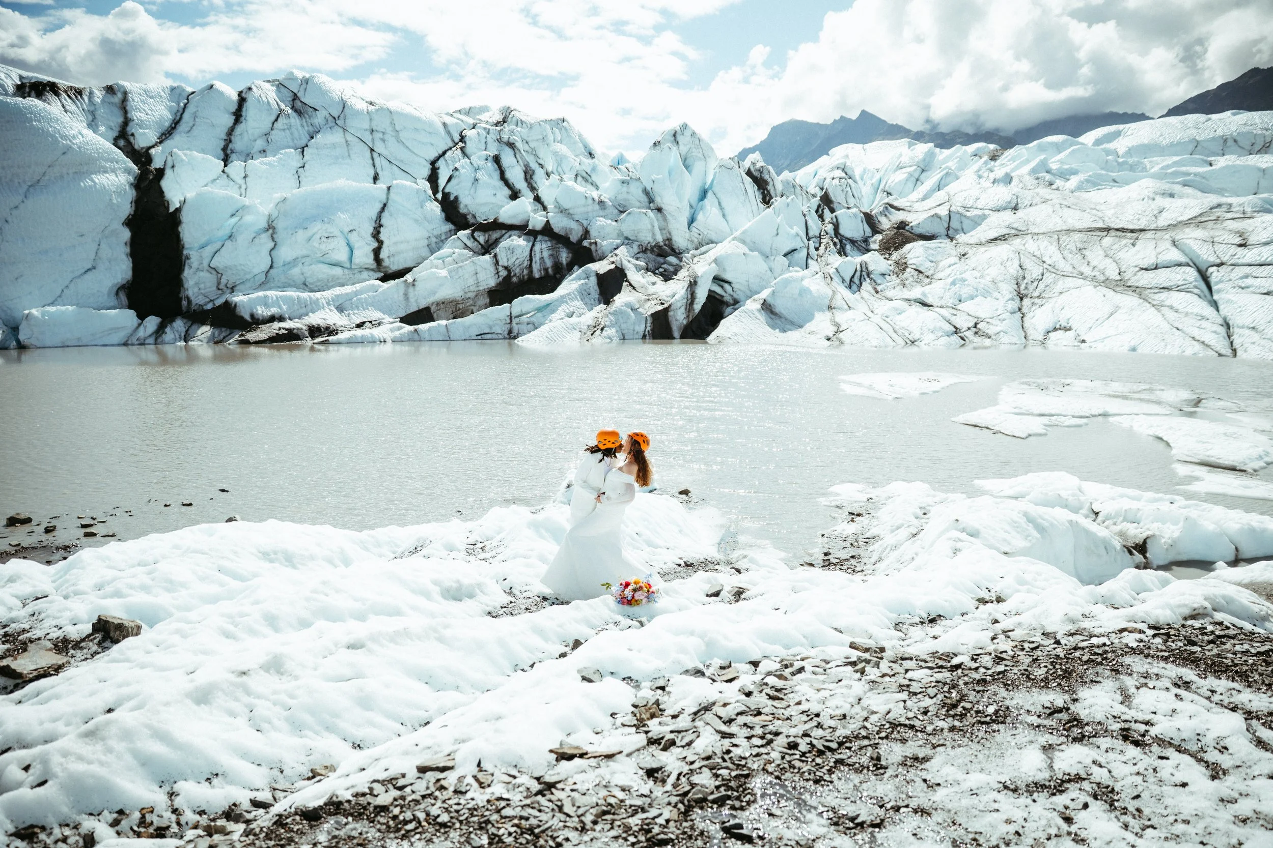 Two women in white wedding dresses and orange helmets stand together on snowy ground near a glacier and icy lake, holding hands.