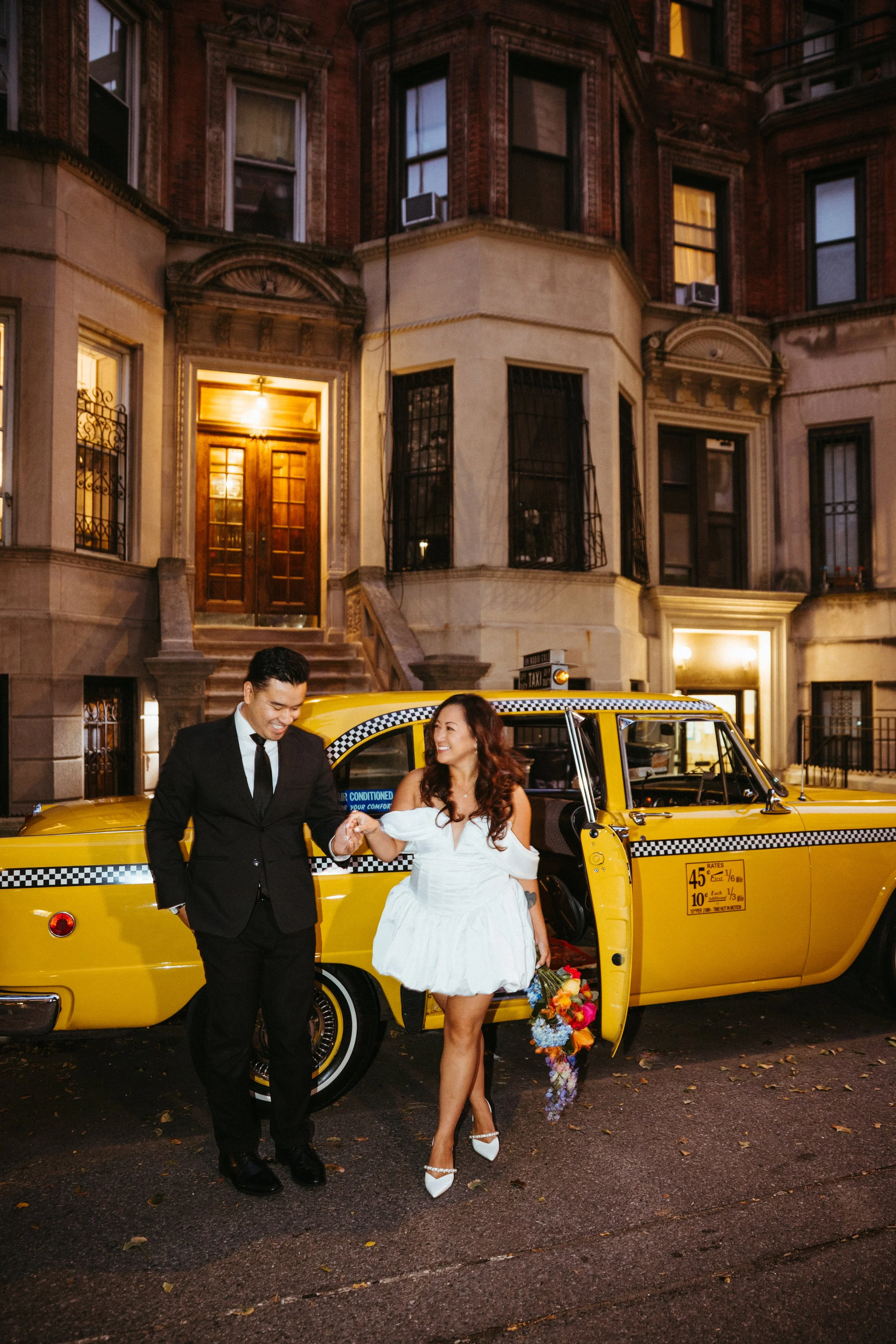 A couple dressed in wedding attire standing outside a yellow taxi cab in front of a brownstone apartment building at night. The groom is wearing a black suit and tie, and the bride is wearing a white dress holding a bouquet of flowers.
