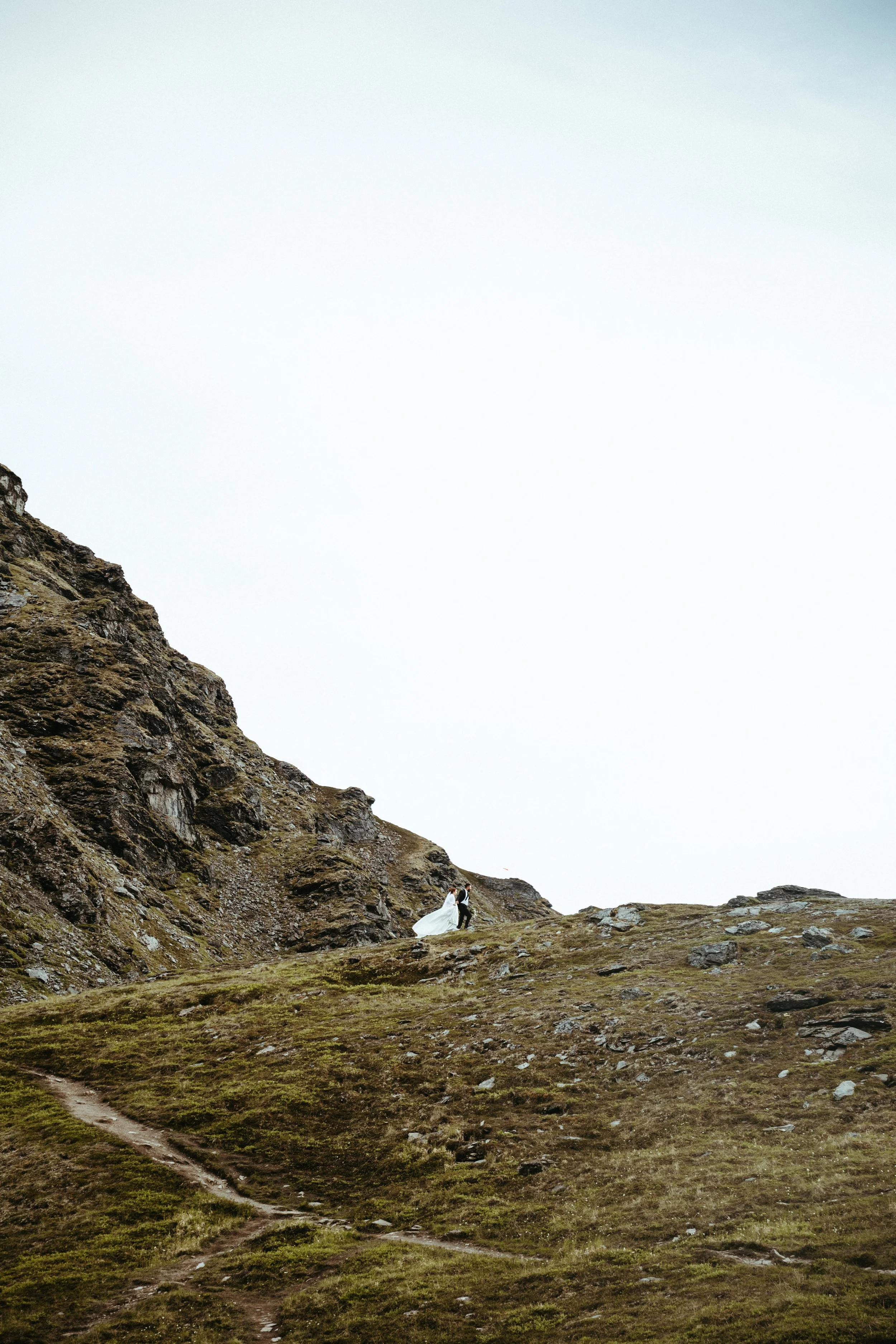 A bride and groom holding hands and walking on a grassy hillside with rocky terrain in the background.