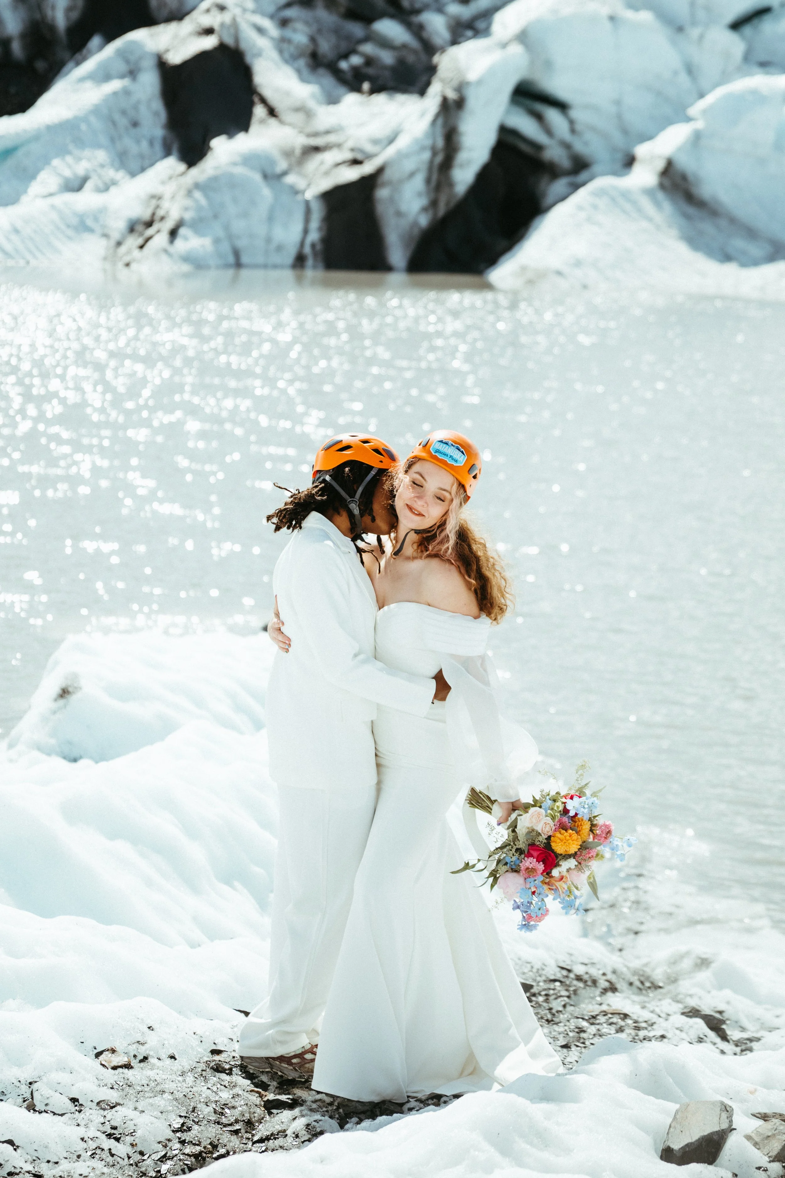A couple in wedding attire standing on snow near a glacier and lake, wearing orange helmets, with one holding a bouquet of colorful flowers.
