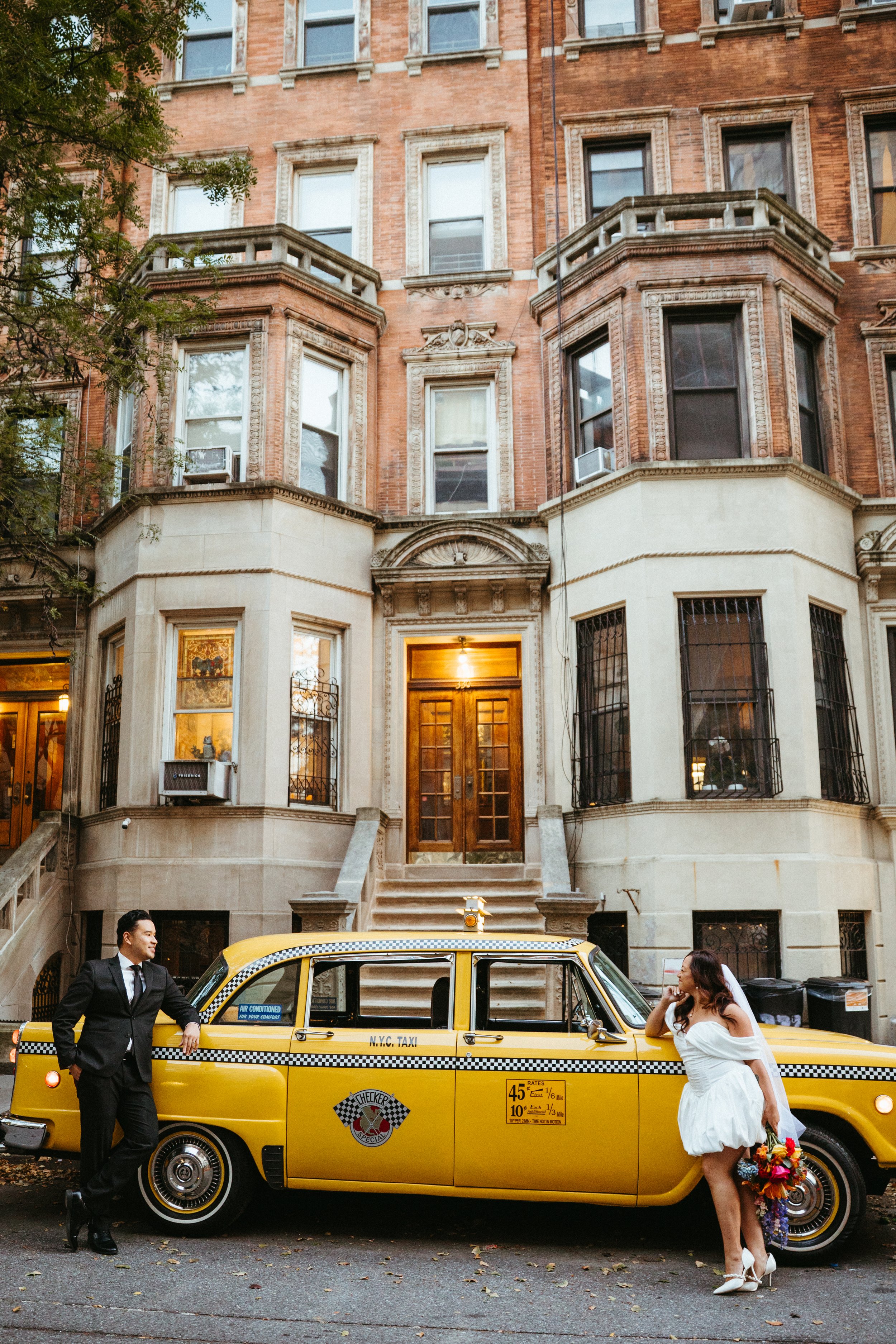 A bride and groom standing beside a yellow NYC taxi cab in front of a brownstone apartment building. The groom is dressed in a black suit, and the bride is wearing a white wedding dress holding a bouquet of colorful flowers.