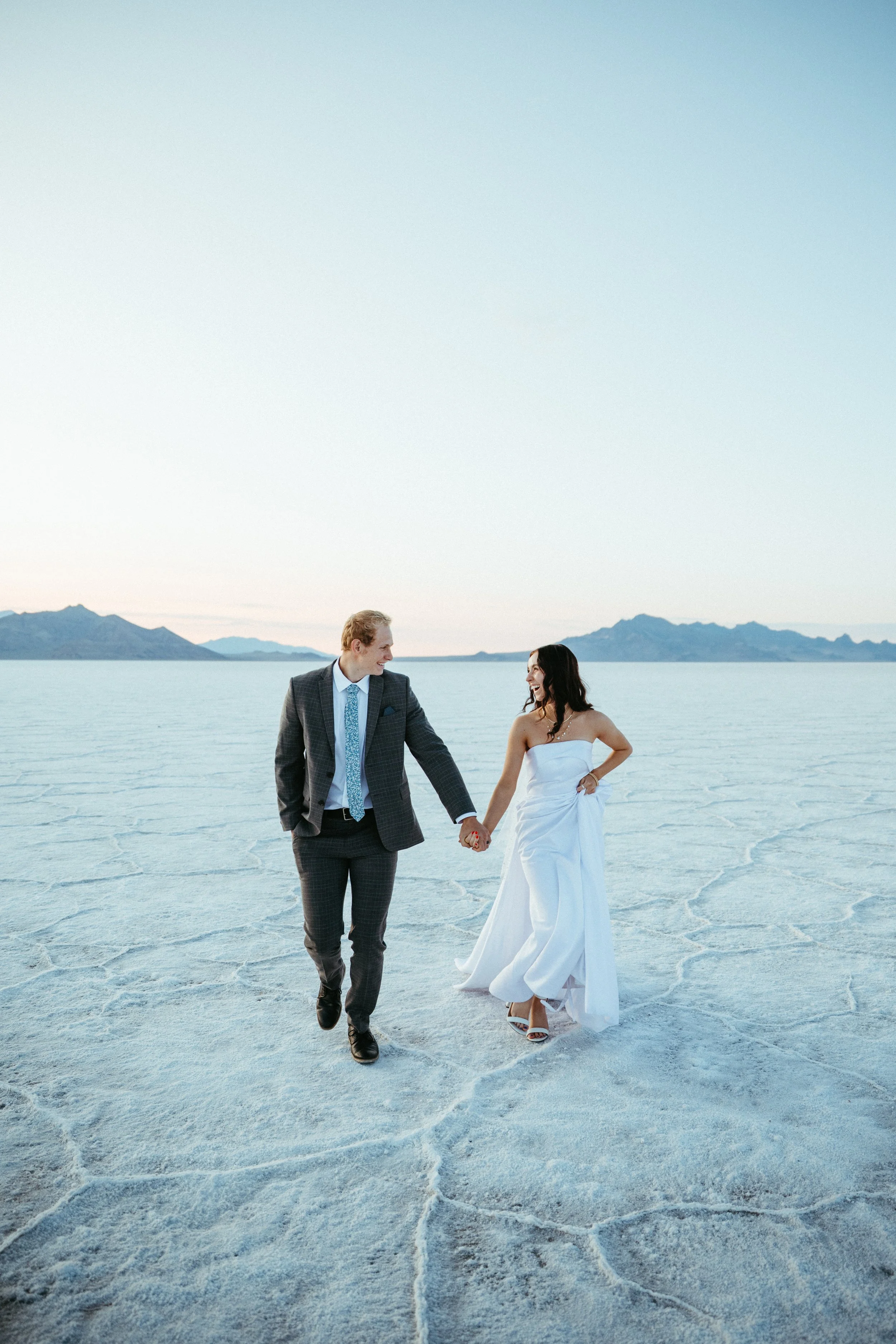 A couple in wedding attire walking hand in hand across a salt flat with distant mountains in the background.