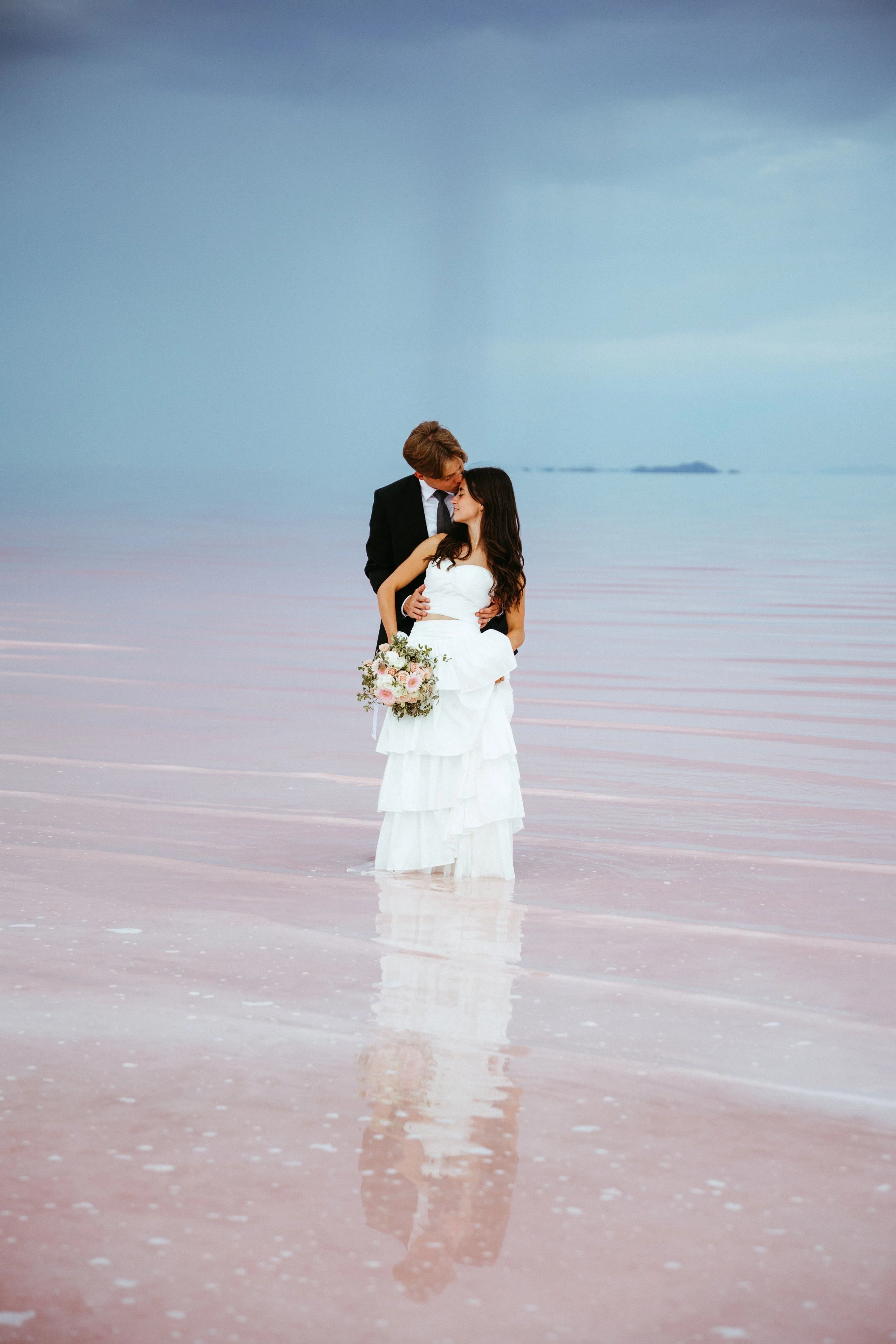 A bride in a white wedding dress and a groom in a black suit are embracing in shallow water on a pink beach, with a distant horizon and cloudy sky in the background.