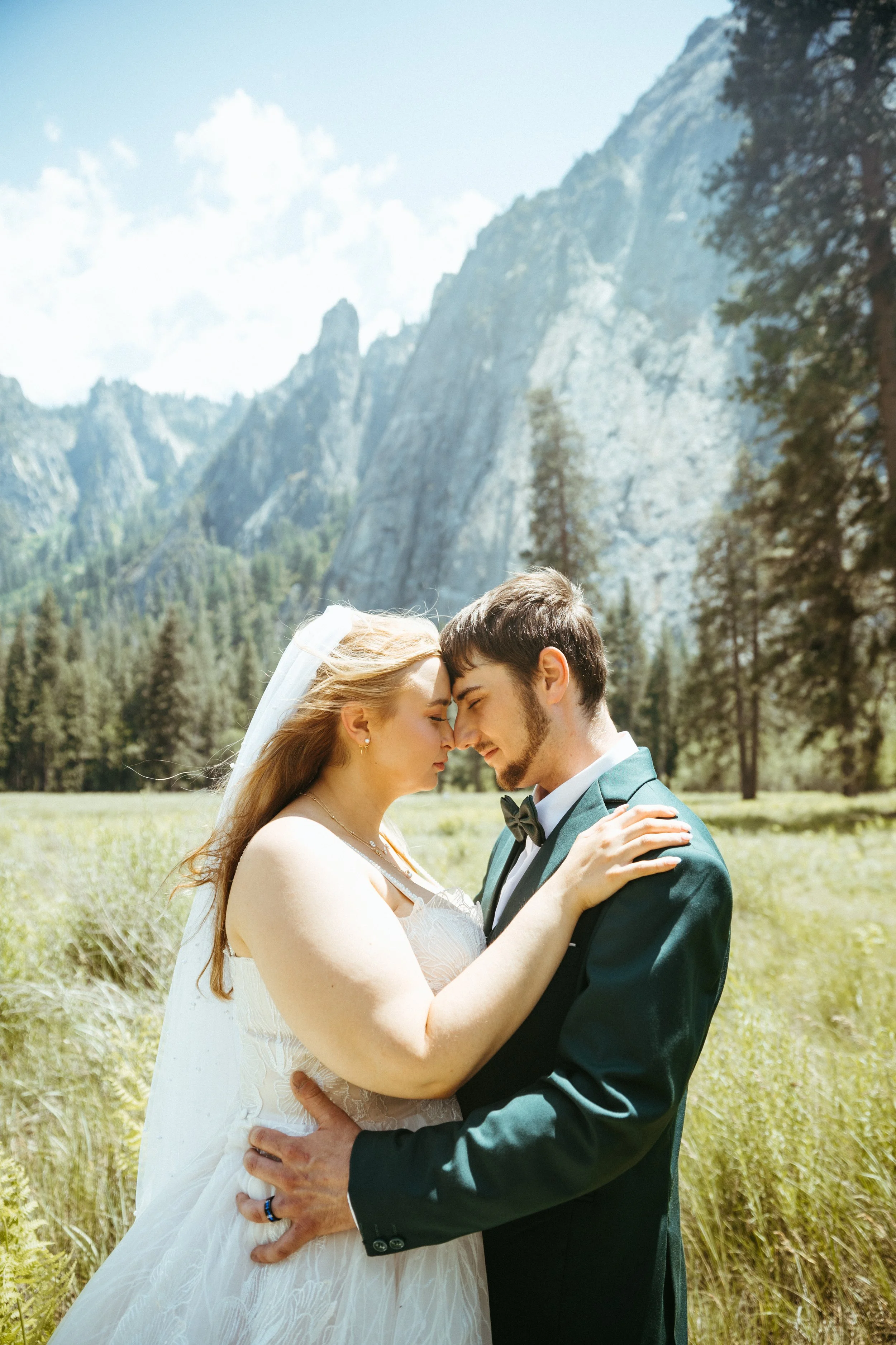 A bride and groom in wedding attire embracing outdoors with mountains and trees in the background.