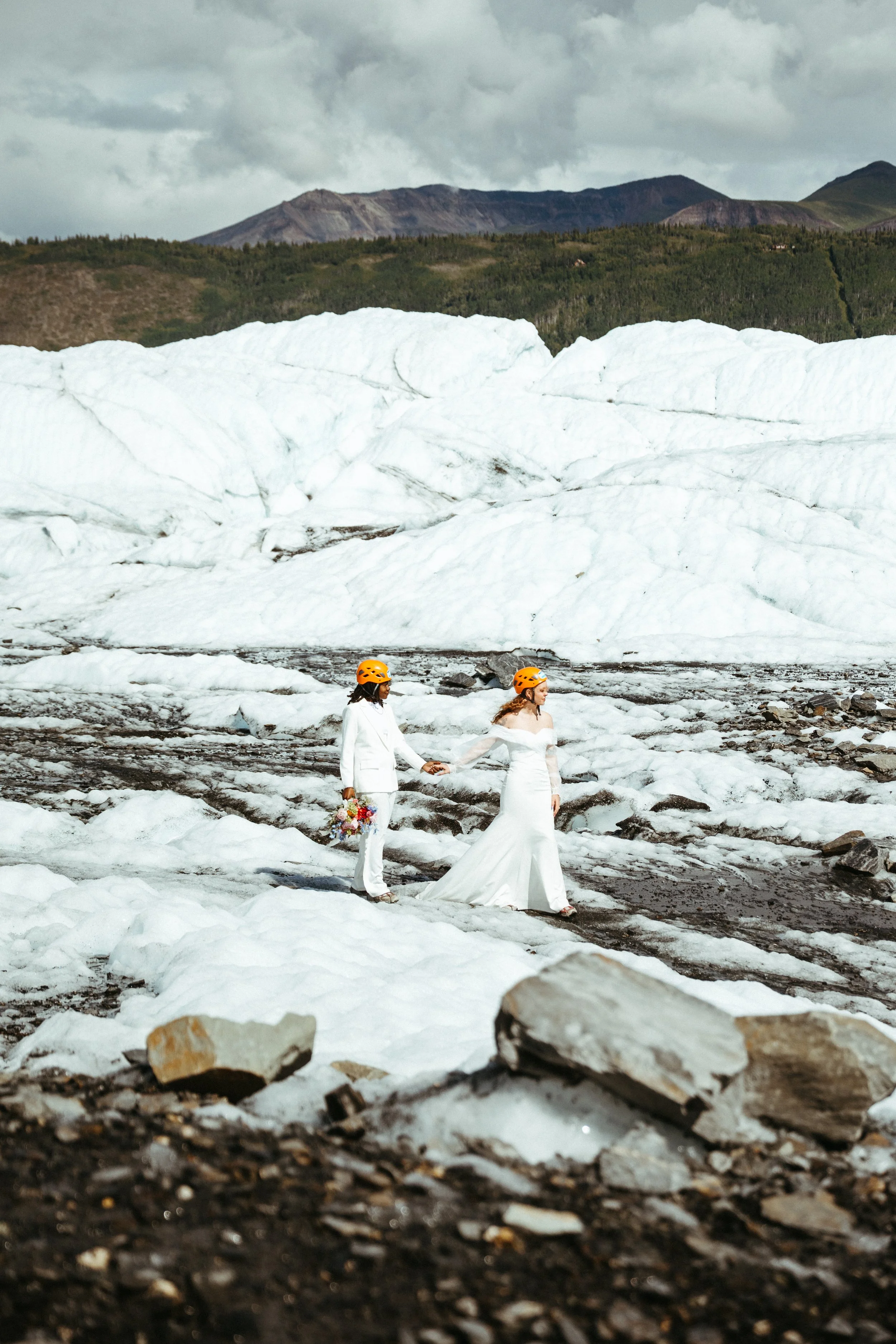 Two women in wedding dresses and orange helmets holding hands on a snowy glacier, with mountains in the background.