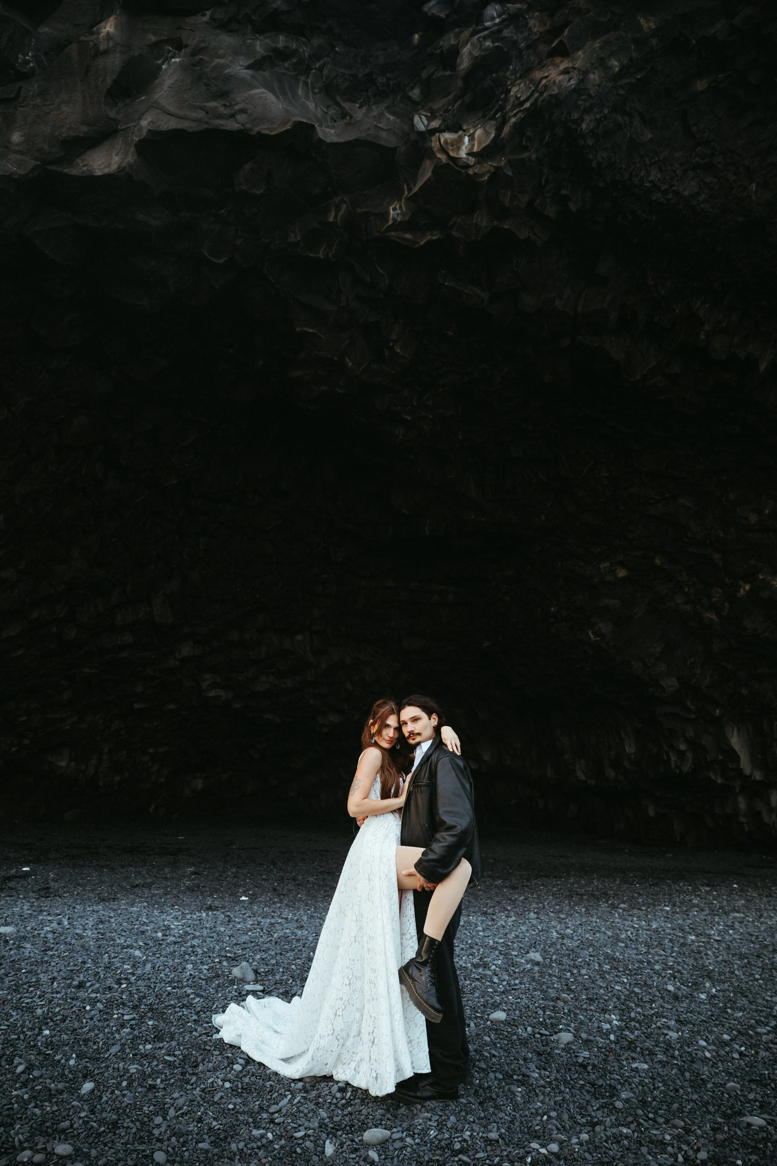 A couple standing on a dark, rocky landscape in front of a large, cave-like rock formation. The woman is wearing a long white dress, and the man is dressed in dark clothing, holding a rolled-up document.