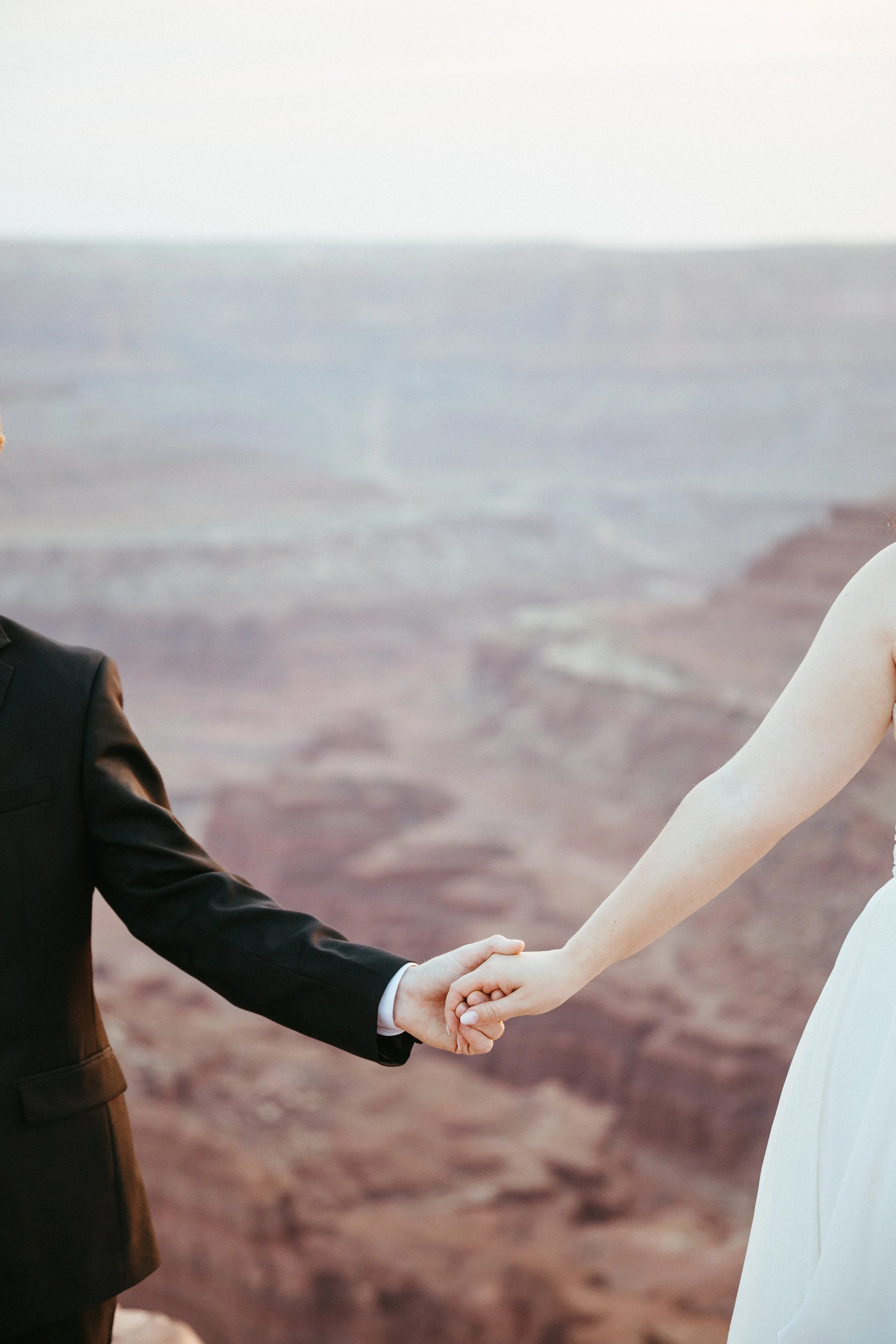A couple holding hands outdoors, with a blurred canyon or rocky landscape in the background. The person on the left is wearing a dark suit, and the person on the right is wearing a white sleeveless dress. Dead horse point state park wedding, Moab