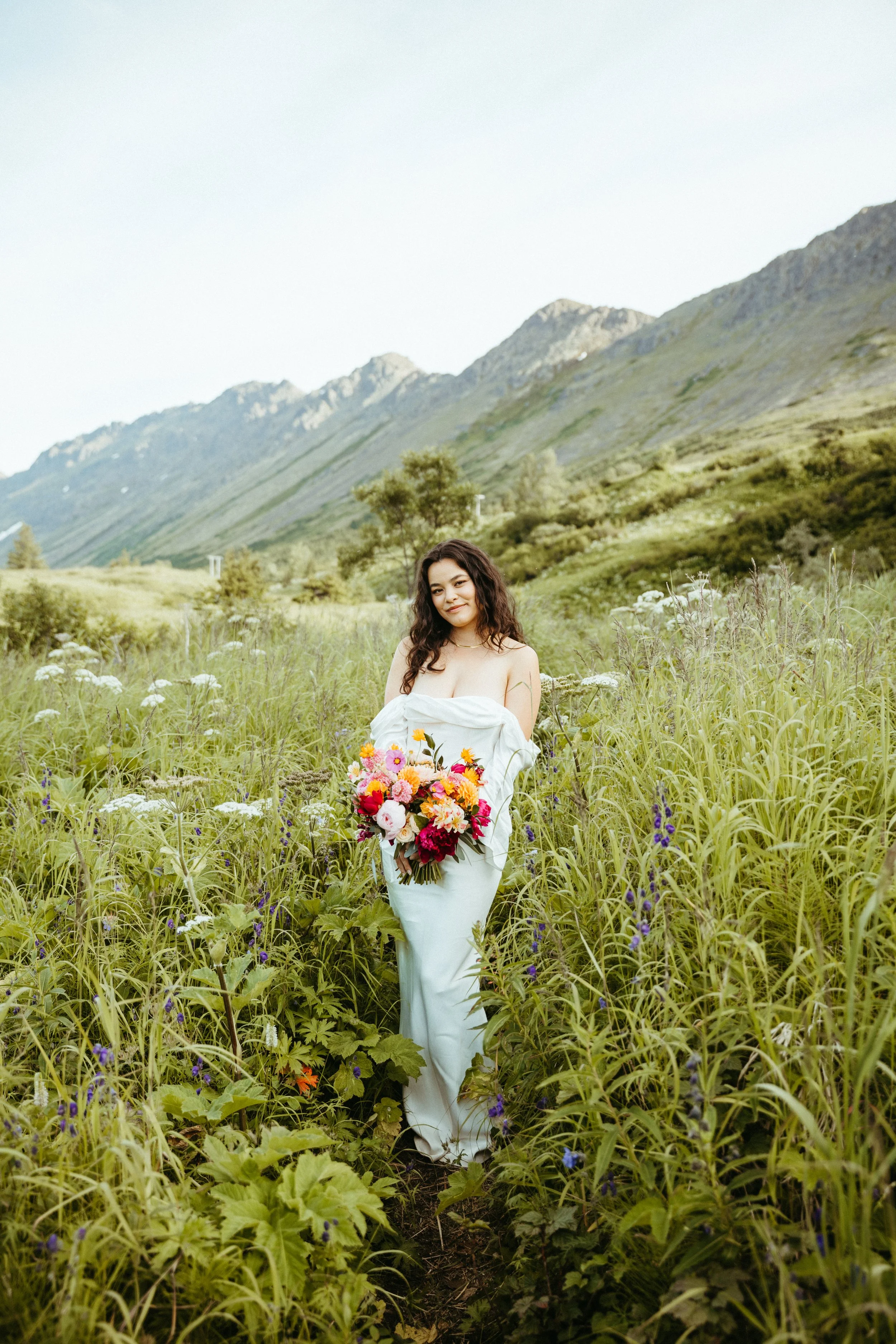 A woman standing in a lush green meadow with mountains in the background, holding a colorful bouquet of flowers, wearing a white dress.