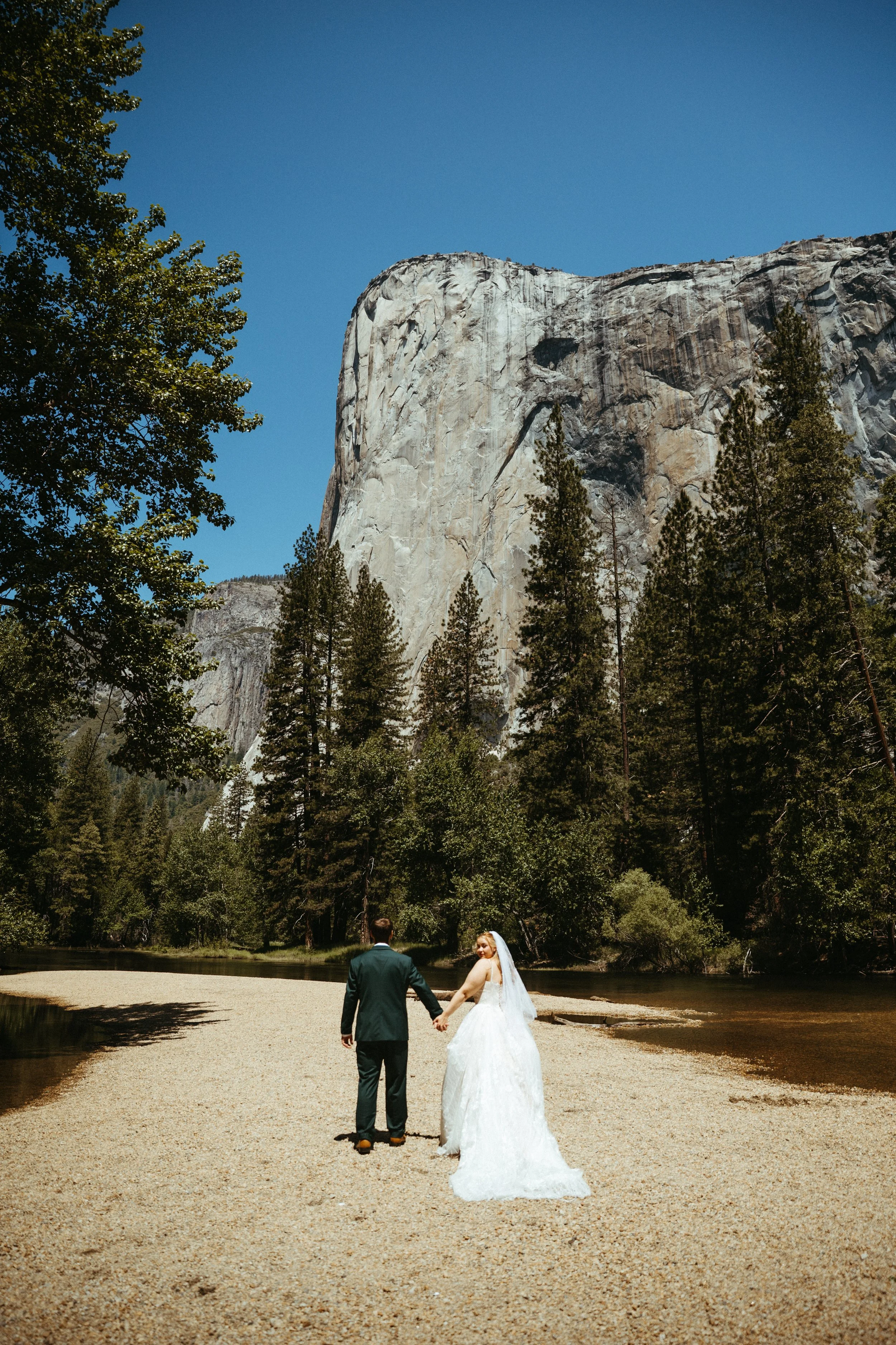 A bride and groom holding hands in a scenic outdoor location with a large rock formation and trees in the background.