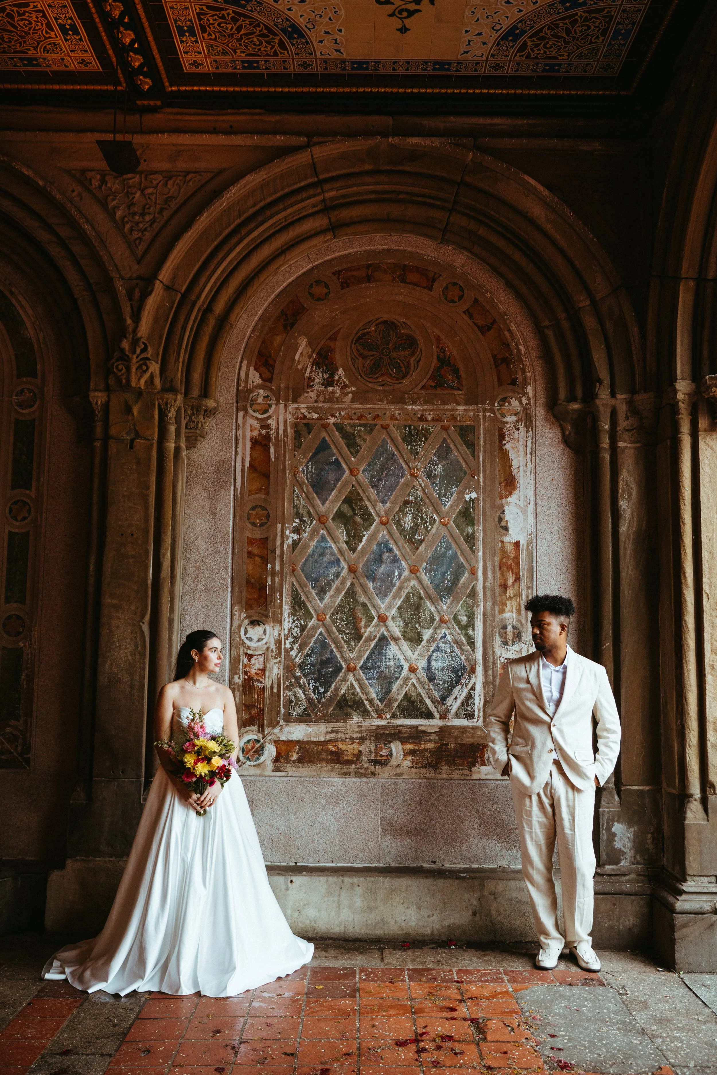 A bride and groom standing apart in front of a historic stone wall with an ornate stained glass window. The bride wears a strapless white wedding gown and holds a bouquet, looking at the groom. The groom wears a cream suit with hands in pockets, faci