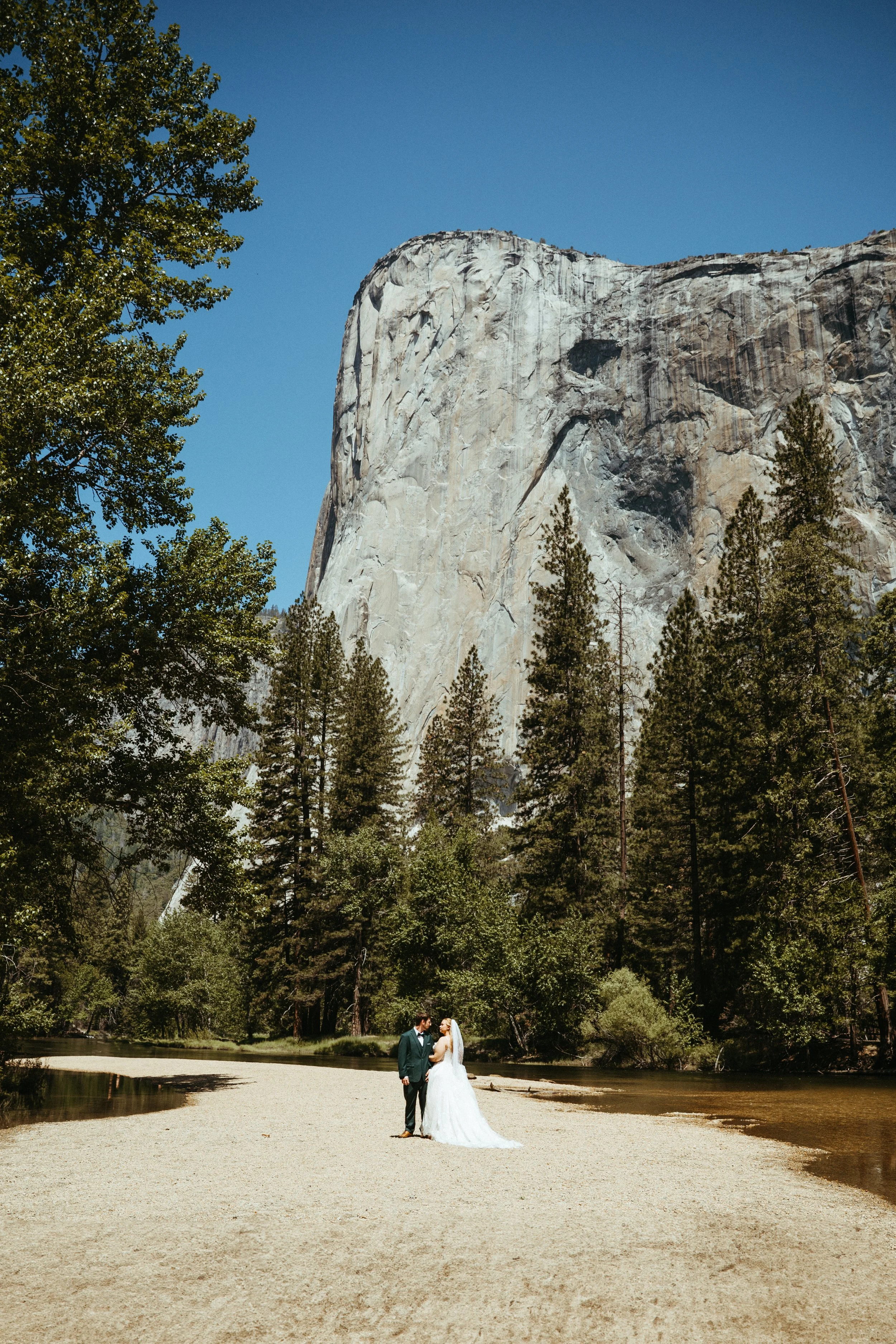 A bride and groom standing on a sandy area near a river, surrounded by tall trees with a large granite cliff in the background under a clear blue sky.