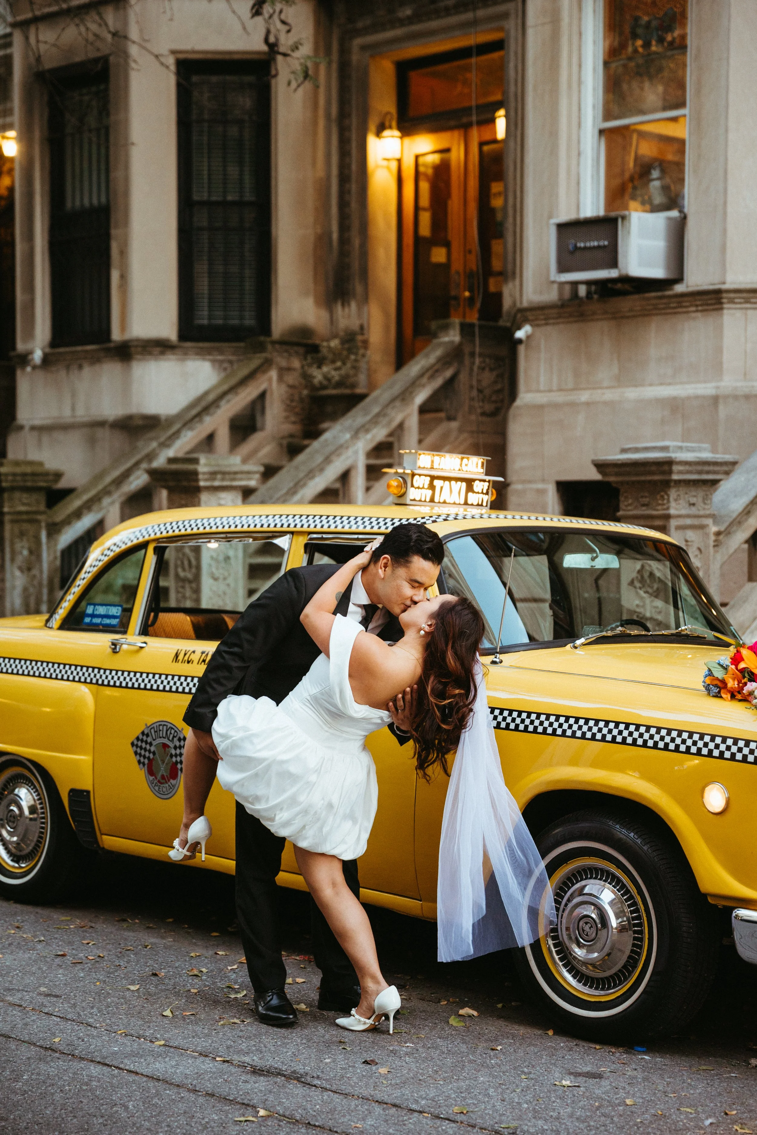 A newlywed couple shares a kiss in front of a yellow taxi cab on a city street at night.