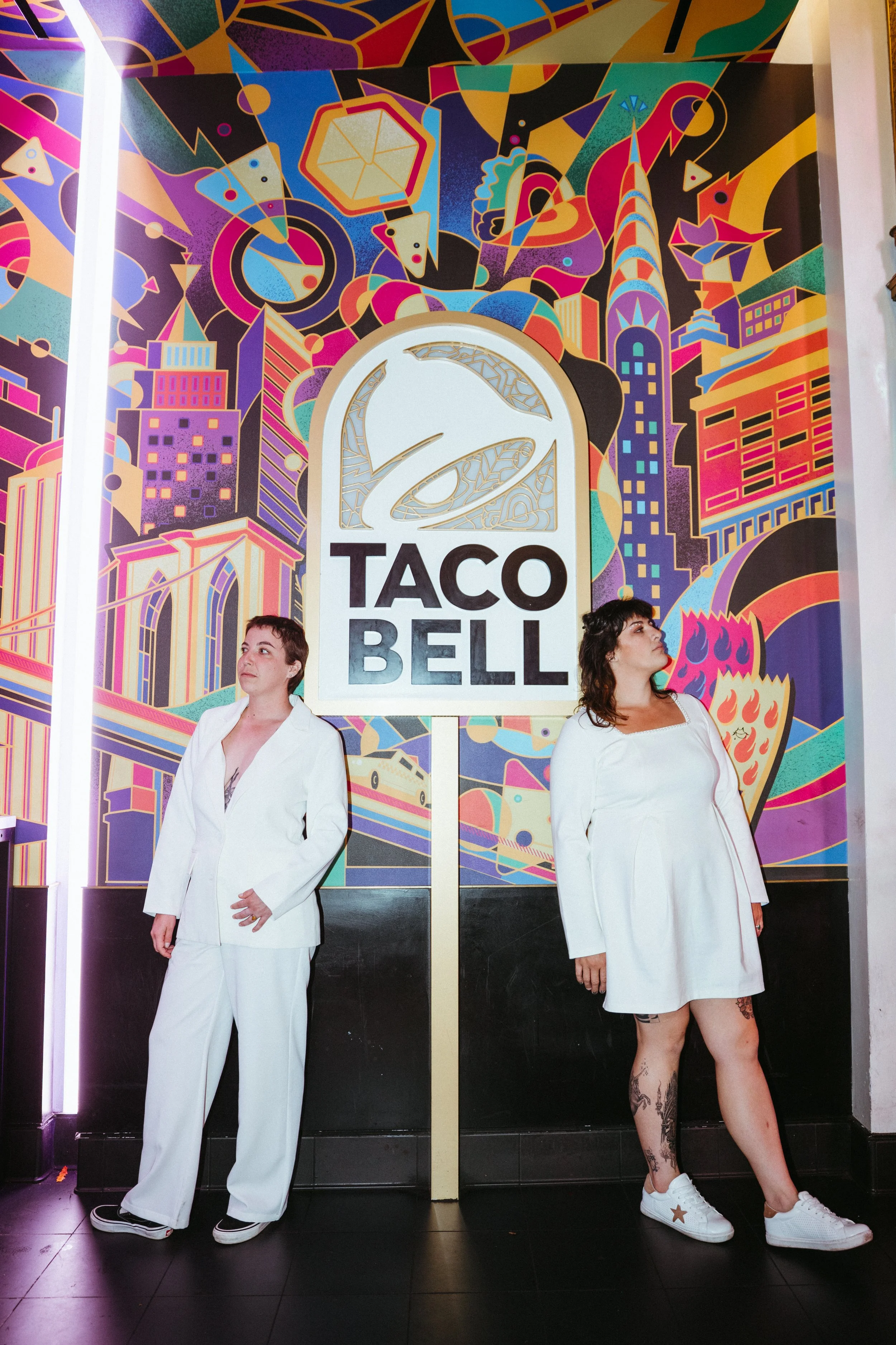 Two women standing in front of a colorful, abstract cityscape mural with a large Taco Bell logo in the center. One woman is wearing white pants and a white blazer, while the other is wearing a white dress and sneakers.