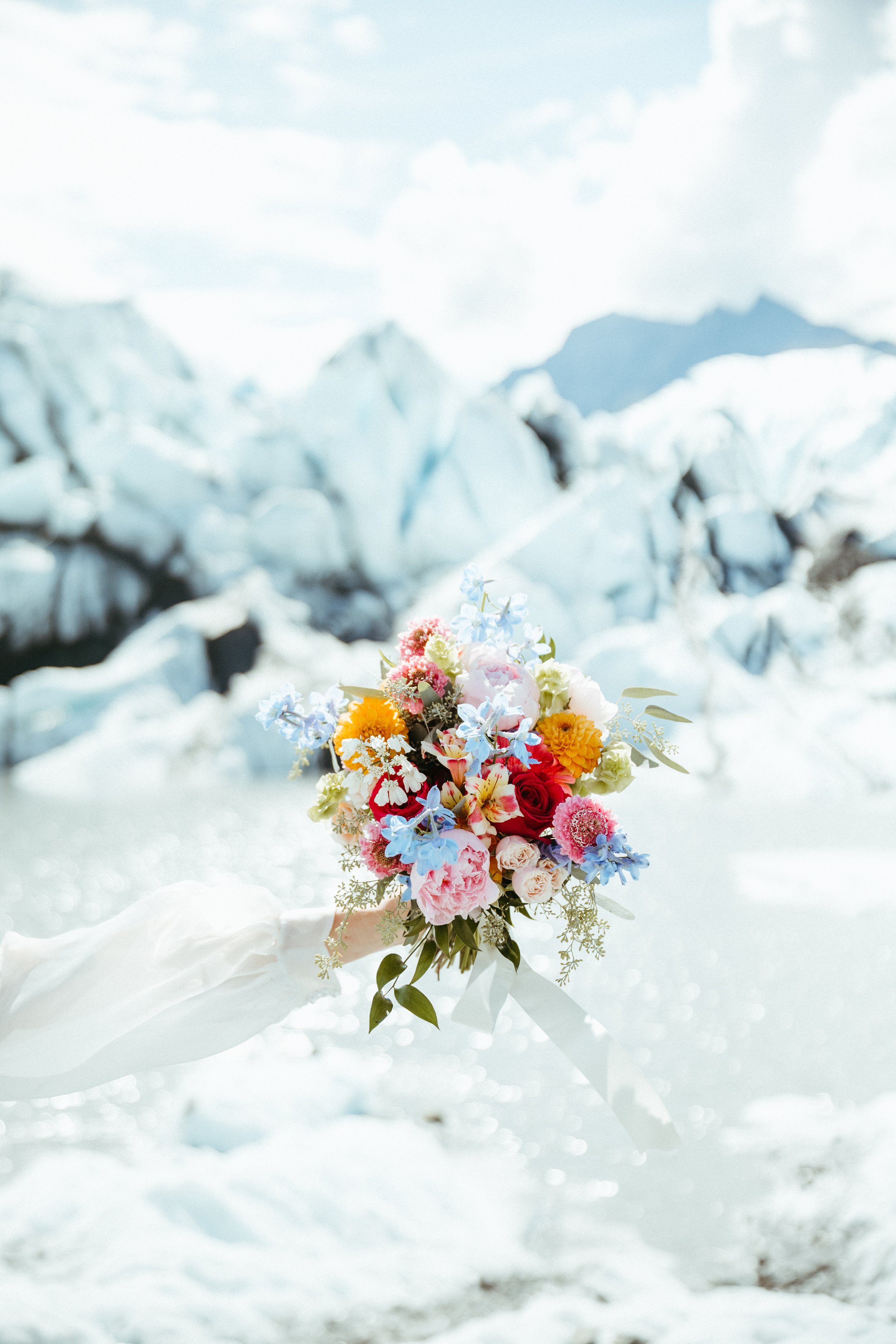 A colorful bouquet of flowers held by a person wearing a white glove, with a snowy mountainous landscape in the background.